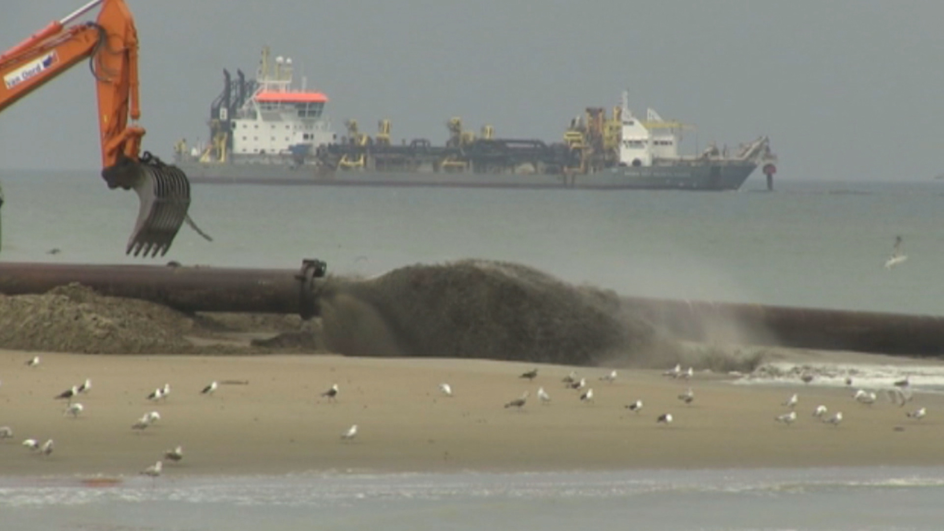 Een baggerschip voert zand aan op het strand via een grote buis, met een graafmachine en meeuwen op de voorgrond.