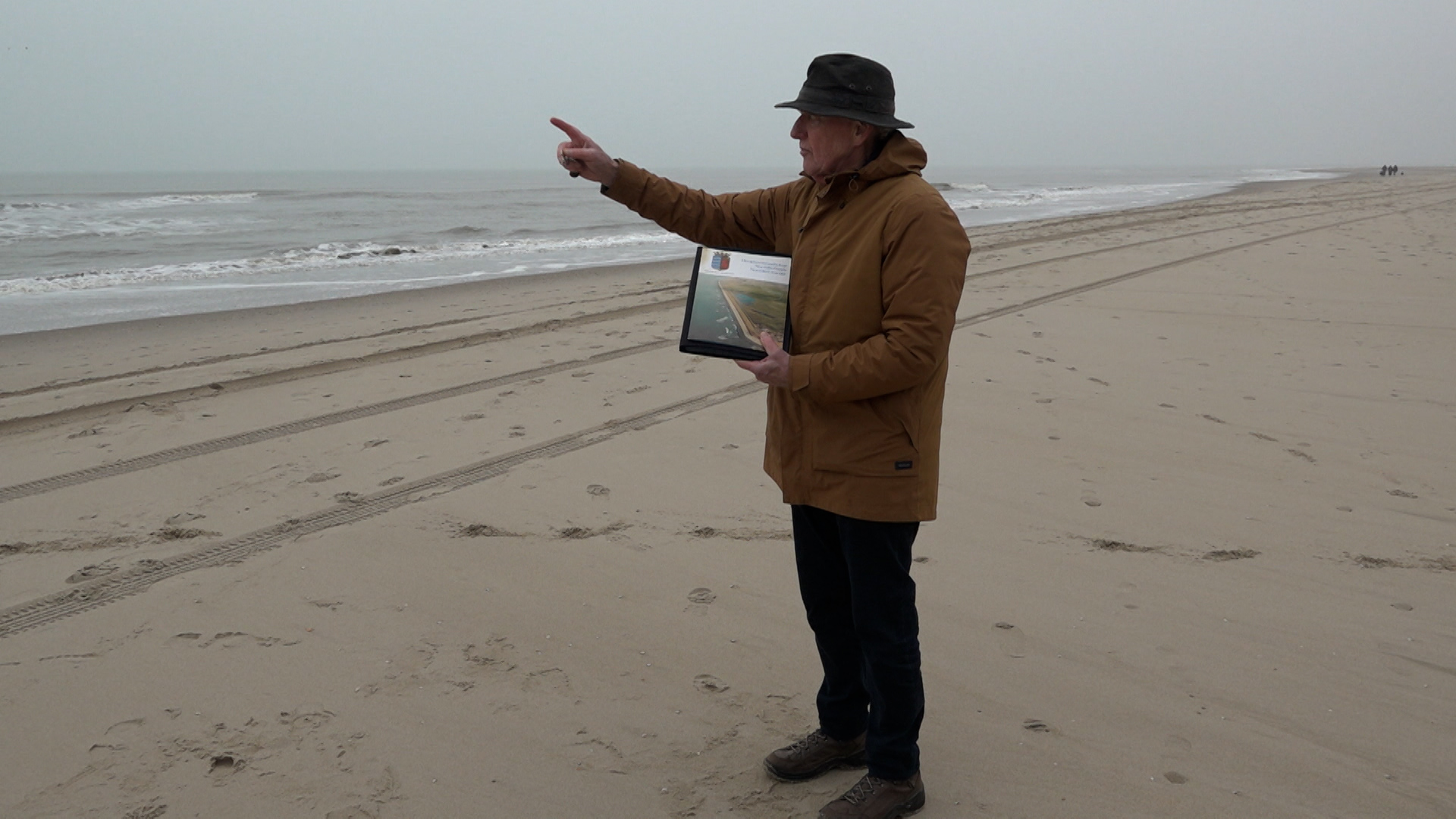 Man in bruine jas wijst naar zee op een leeg strand, met een map in zijn hand.