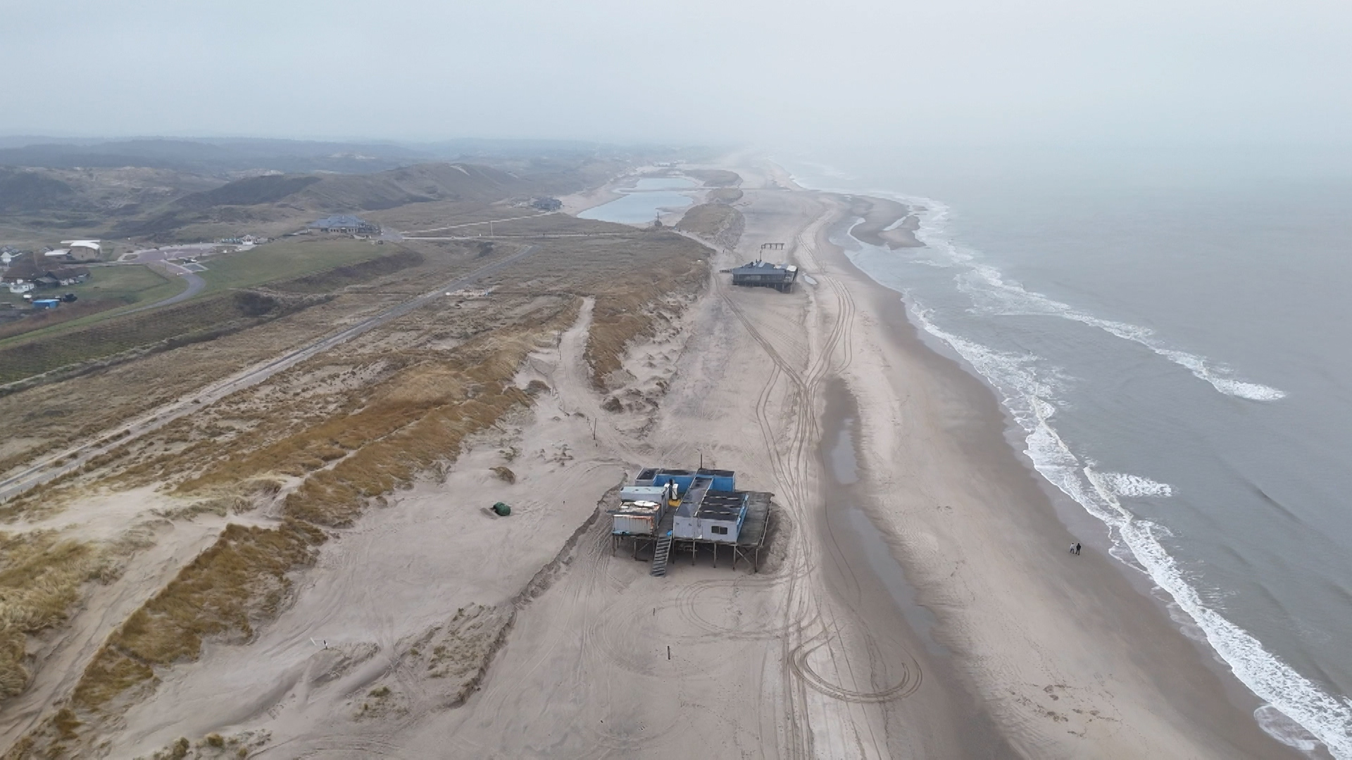 Strandlandschap met zandduinen, enkele strandpaviljoens op palen en de zee aan de rechterkant.