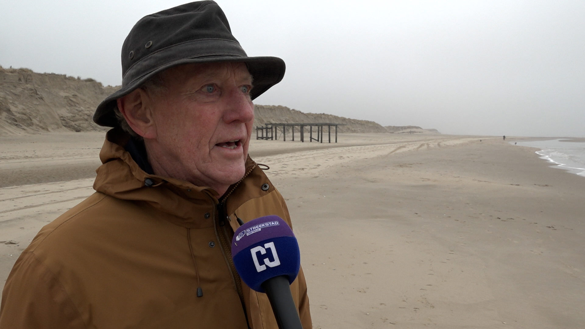 Oudere man met hoed wordt geïnterviewd op een leeg strand met een microfoon met logo van Streekstad, onder een grijze hemel.