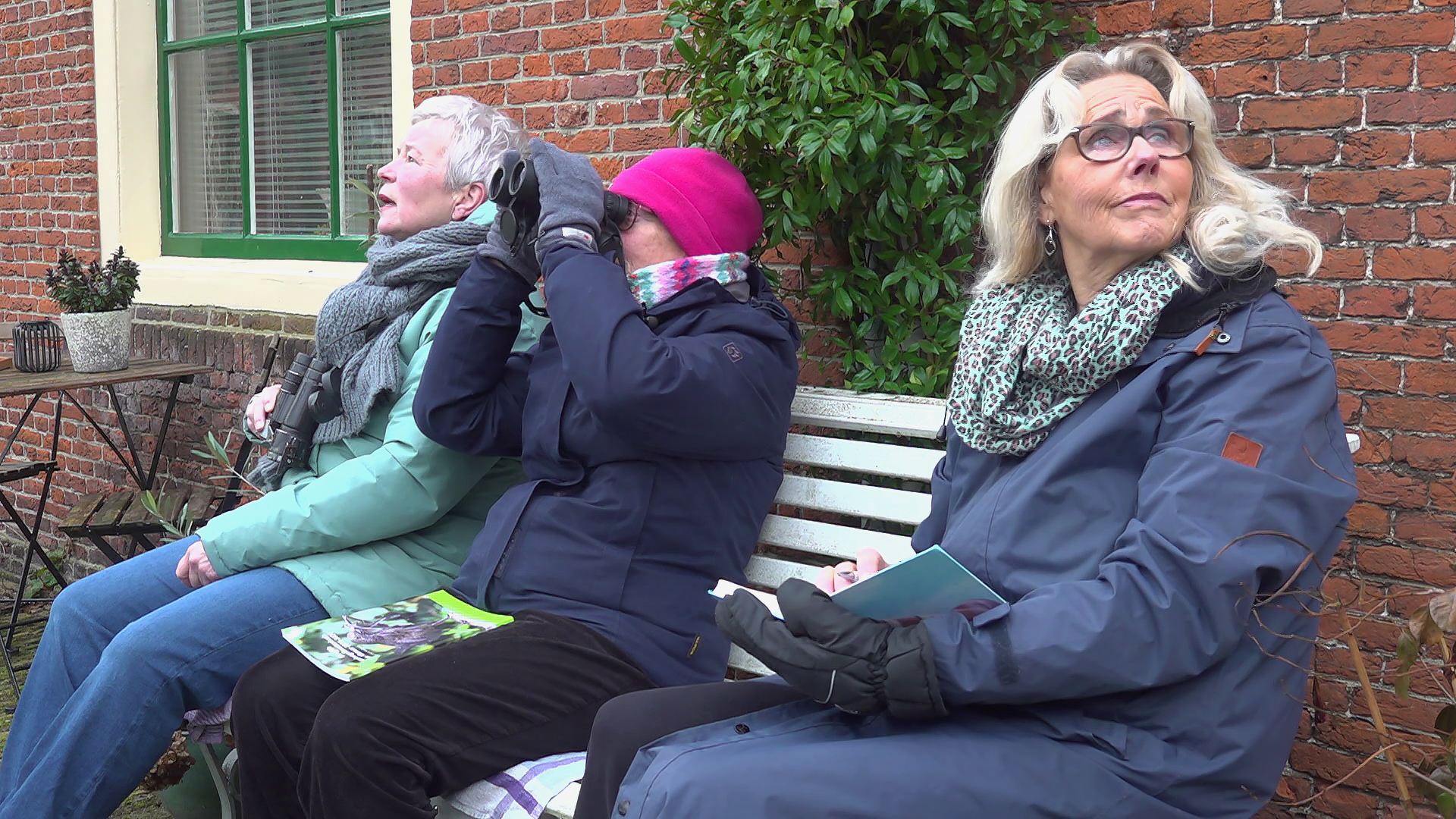 Drie oudere vrouwen zitten op een bank buiten, waarbij één door een verrekijker kijkt en de anderen naar de lucht staren.
