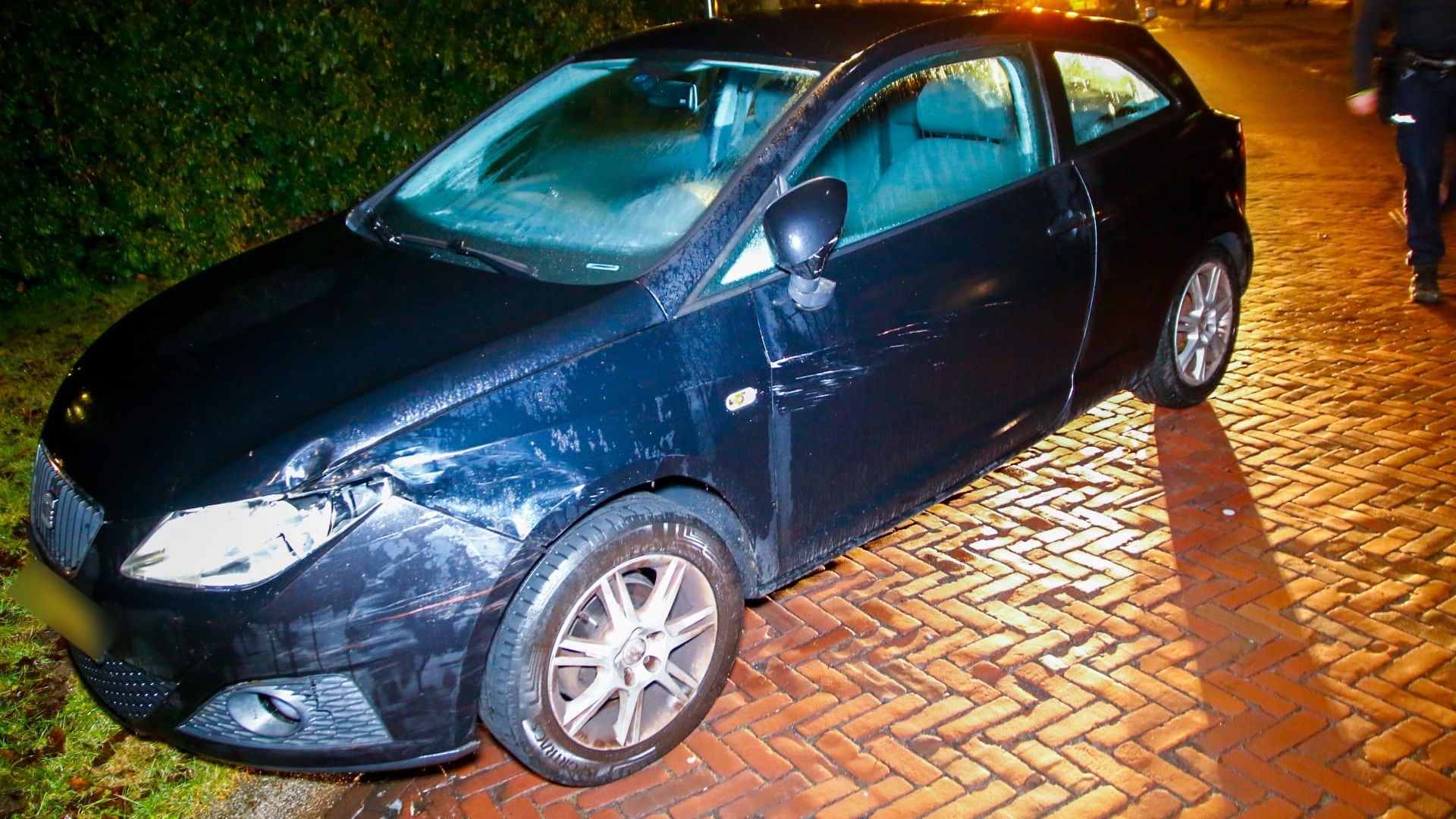 Beschadigde zwarte auto met krassen en deuken aan de voorkant, geparkeerd op een natte bakstenen weg.