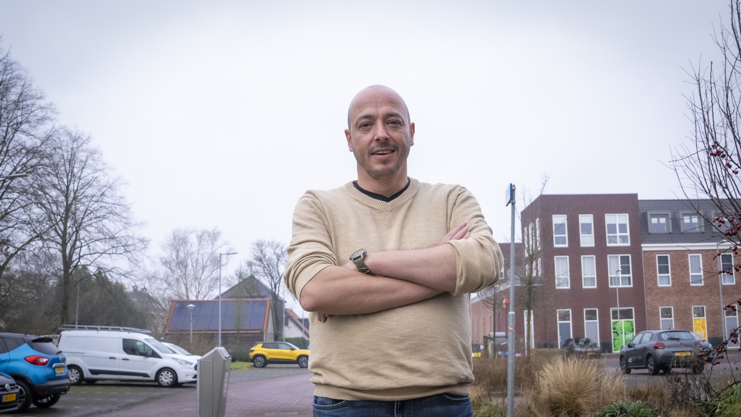 Man in een beige trui met gekruiste armen staat buiten voor een gebouw en geparkeerde auto's.