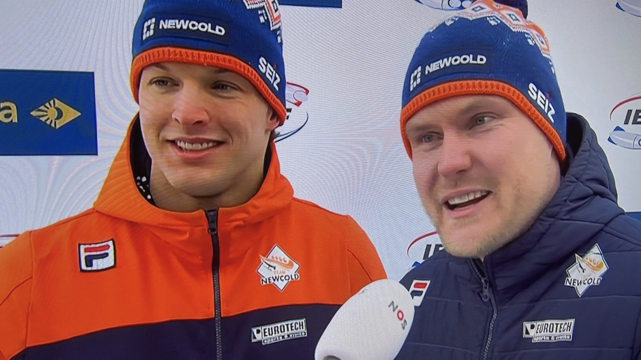 Twee mannen in oranje en blauwe sportkleding geven een interview, er is een microfoon met NOS-logo zichtbaar.