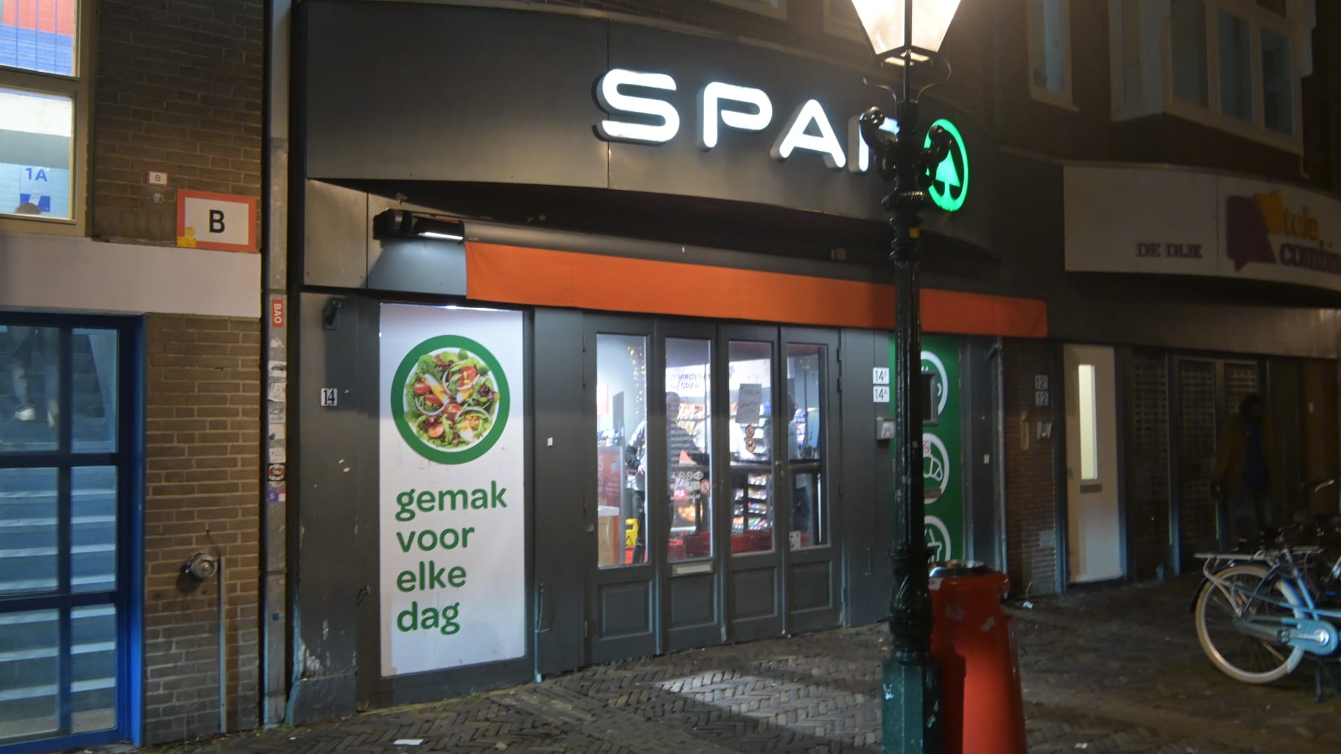 De ingang van een SPAR-winkel in de avond, met een verlichte buitenlamp en fietsen geparkeerd voor de deur.