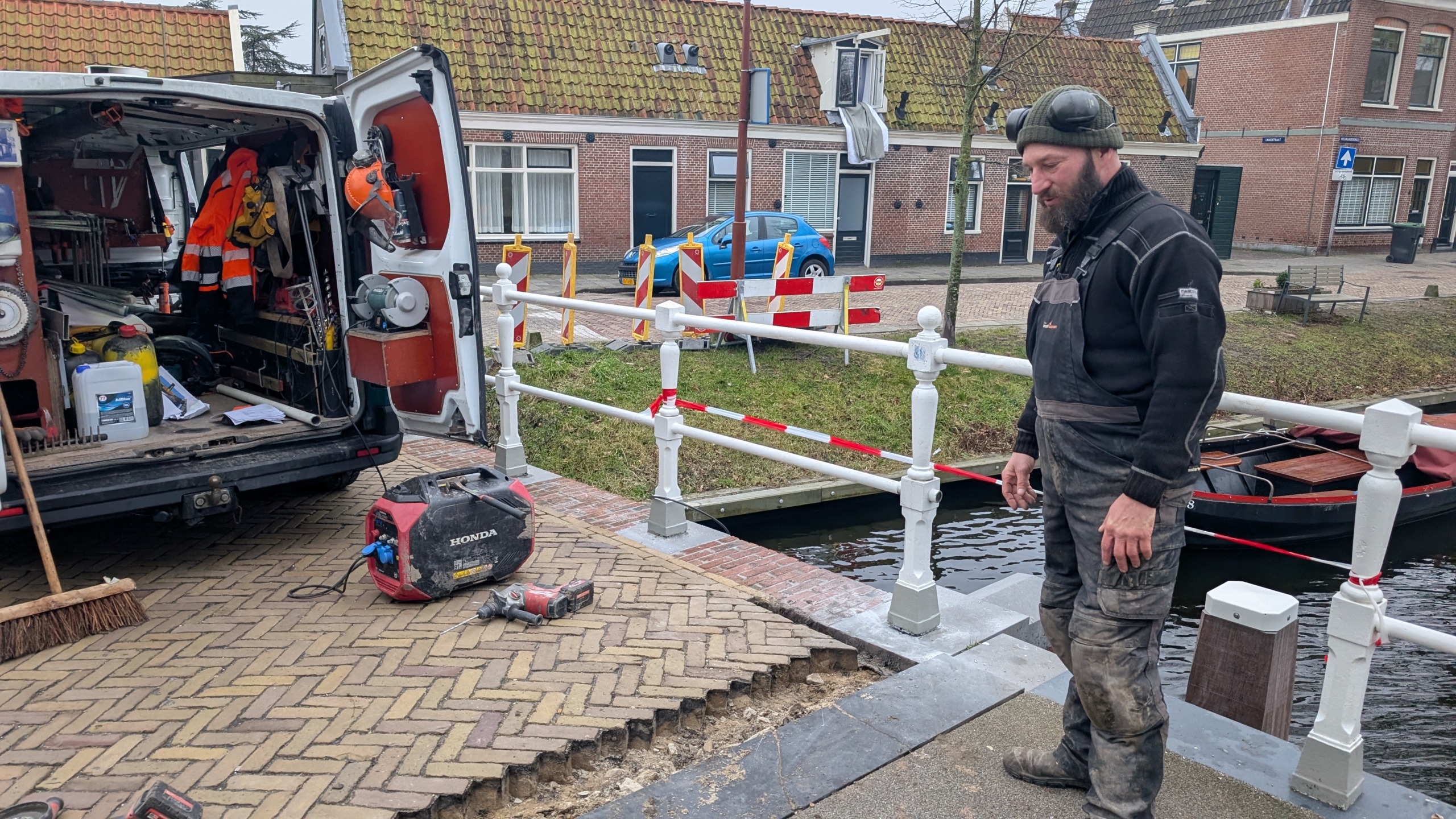 Werkman in outdoor kleding staat naast een busje vol gereedschap bij een brug, met een deels opgebroken bestrating en een sloot op de achtergrond.