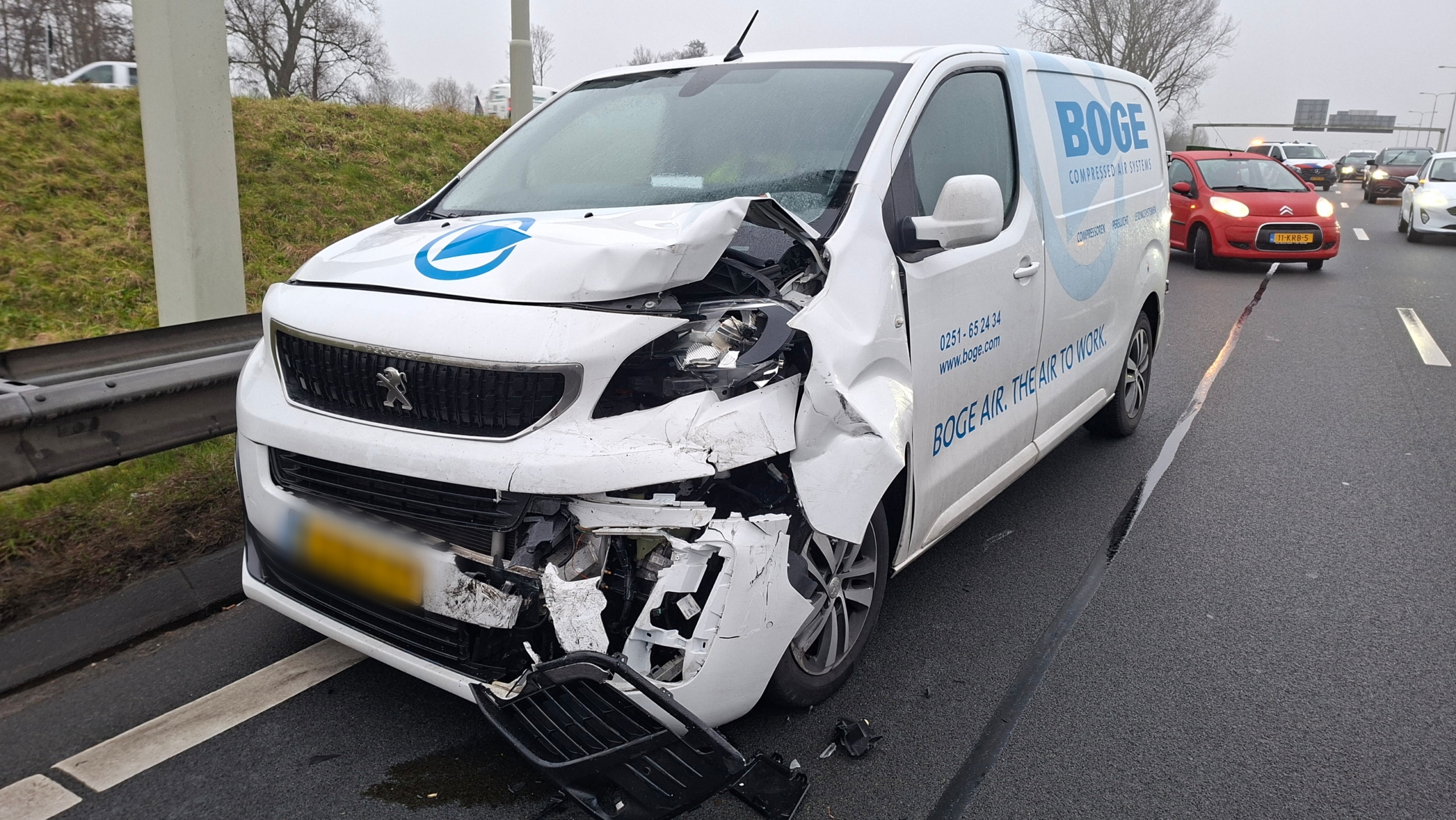 Een witte bestelwagen met zware schade aan de voorkant staat langs de weg na een ongeval.