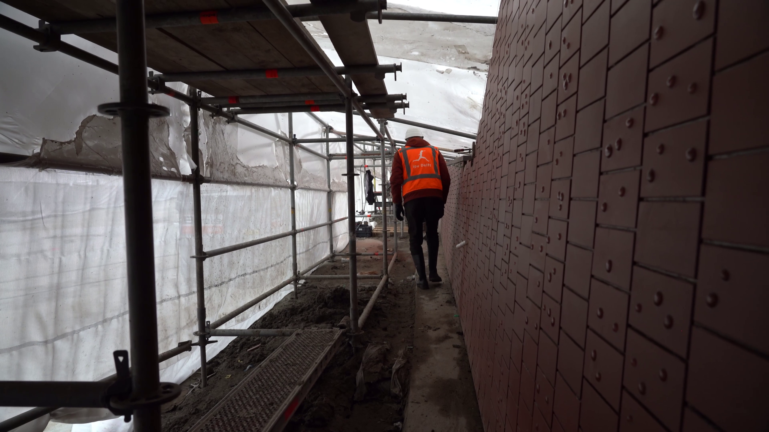 Persoon in oranje vest loopt onder steigervloer langs een muur met donkerrode tegels.