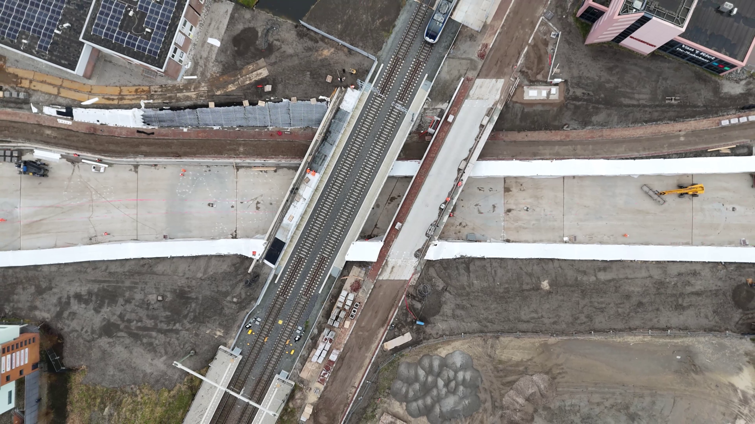 Bovenaanzicht van een bouwplaats met een spoorlijn door een brug, omringd door gebouwen en machines.