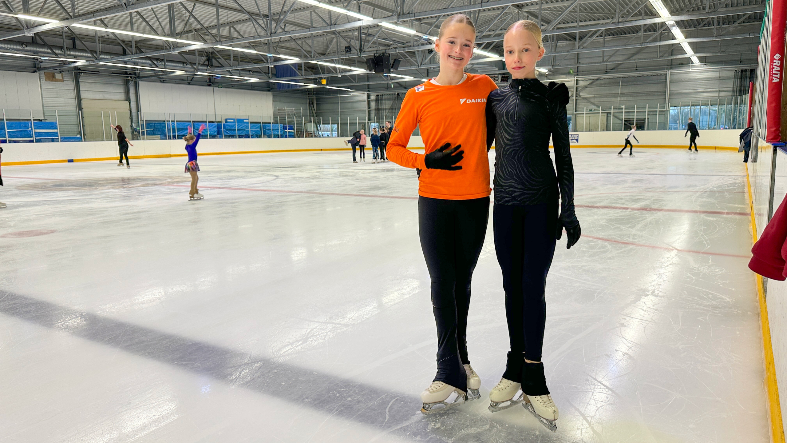 Twee jonge schaatsers poseren op een ijsbaan, omgeven door andere schaatsers in beweging.