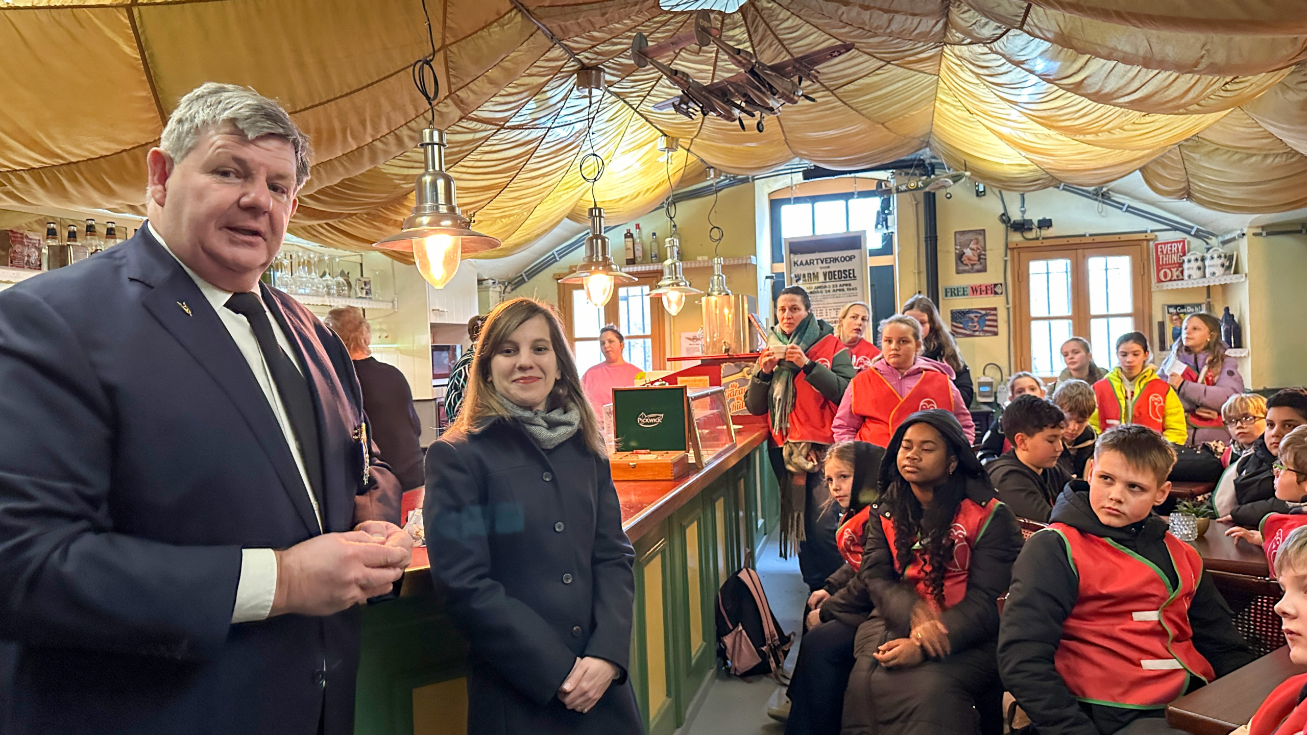 Een groep mensen en kinderen, sommigen in rode truien, luistert naar een man in een pak in een café-achtige omgeving.