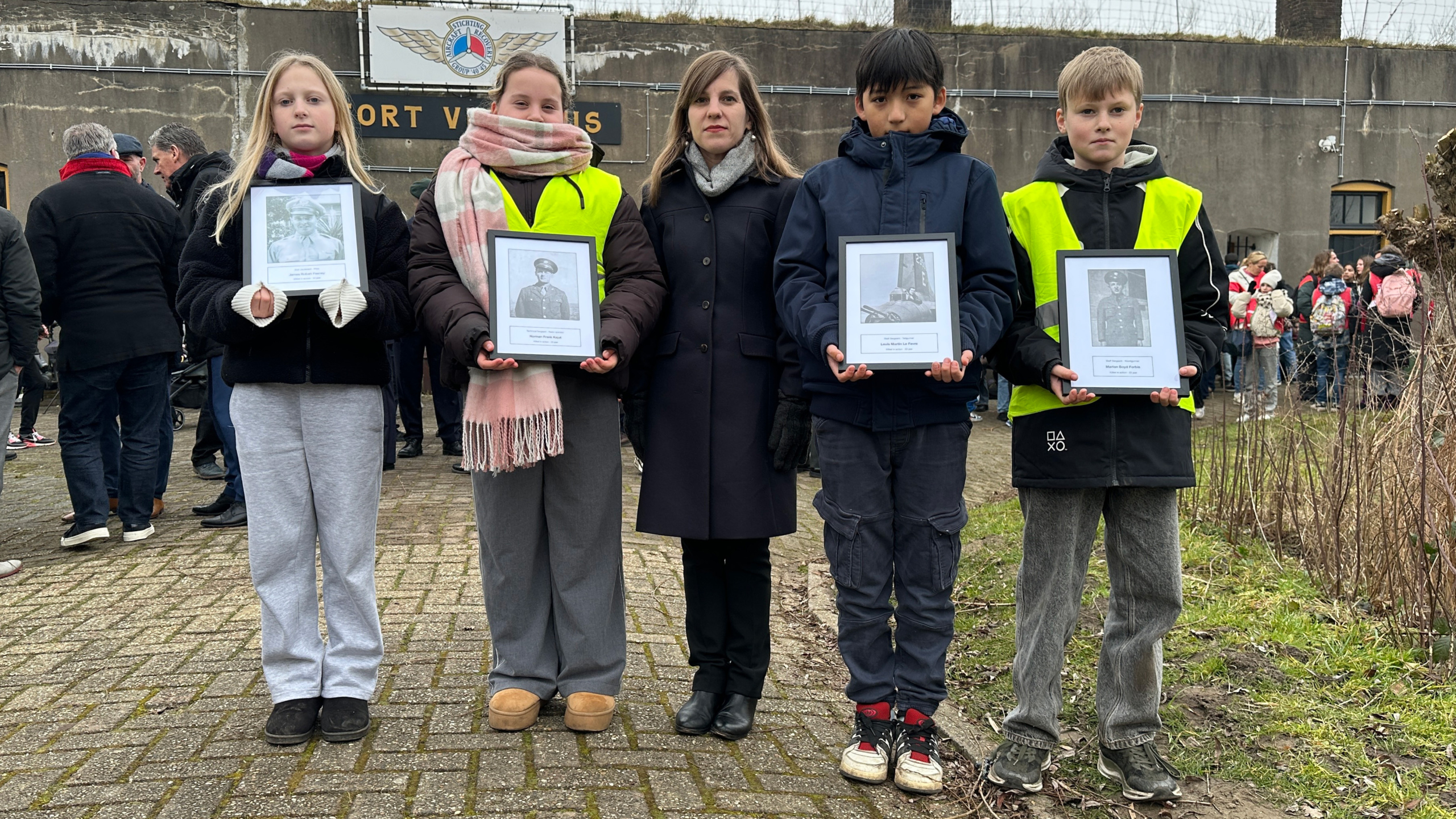 Een groep kinderen en een volwassene houdt ingelijste foto's vast tijdens een ceremonie bij Fort Vijfhuizen, met mensen op de achtergrond.