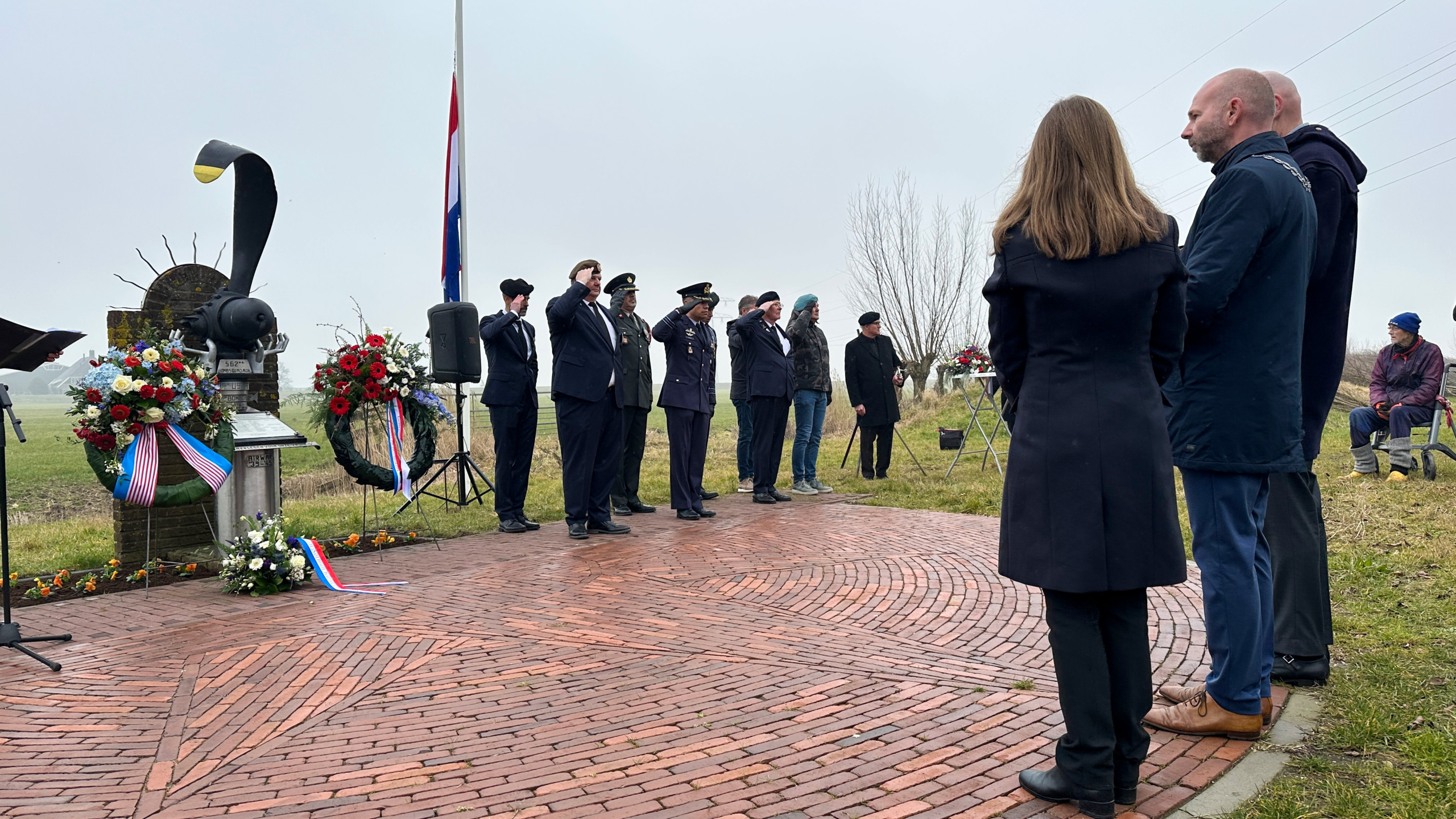 Mensen in uniform wonen een herdenking bij bij een monument, omringd door kransen en vlaggen.