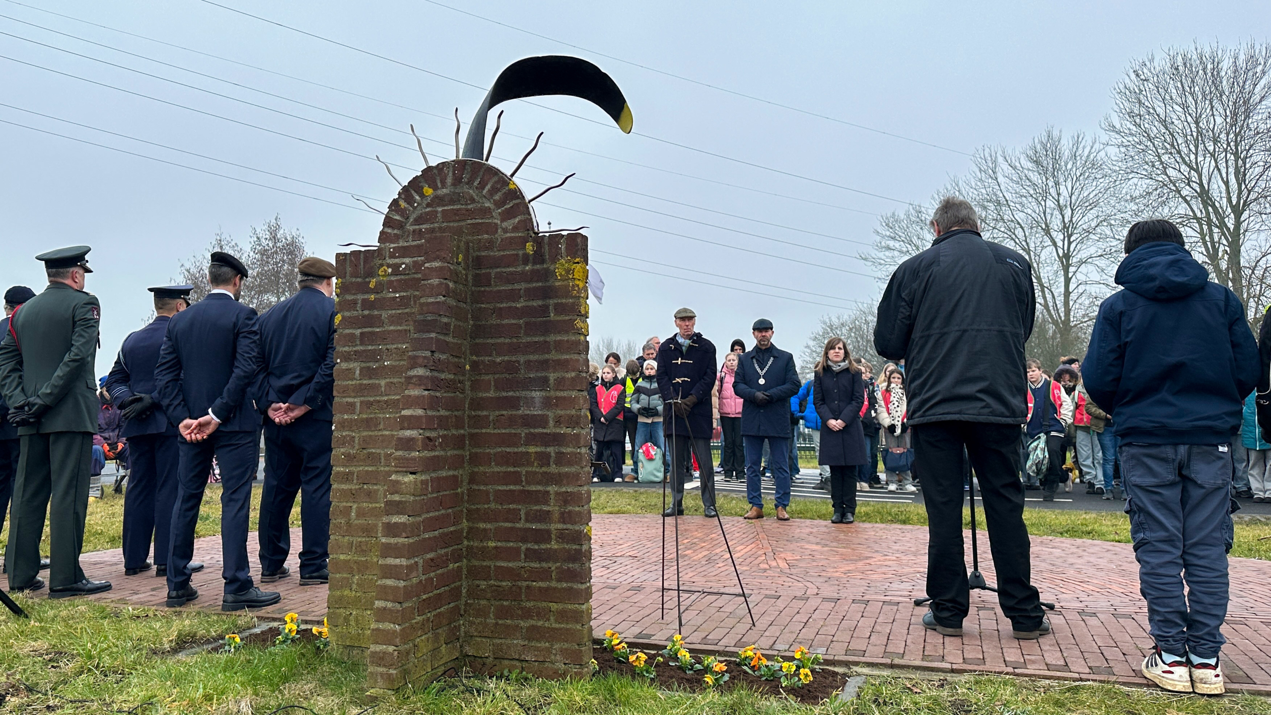 Mensen verzamelen zich voor een ceremonie bij een bakstenen monument met een herdenkingskrans, met bloemen op de grond.