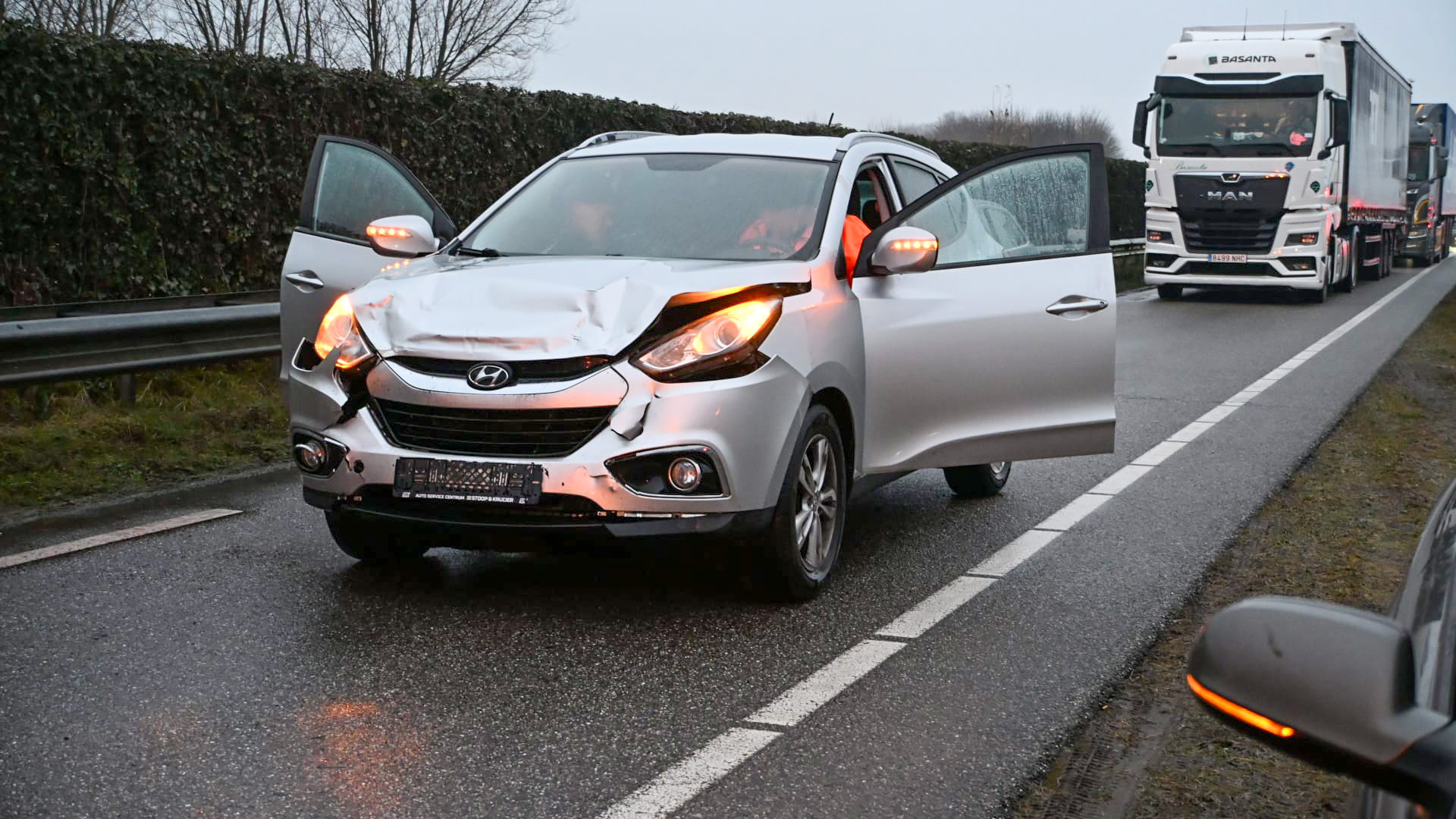 Auto met schade aan de voorkant op een weg, met open portieren en een vrachtwagen op de achtergrond.