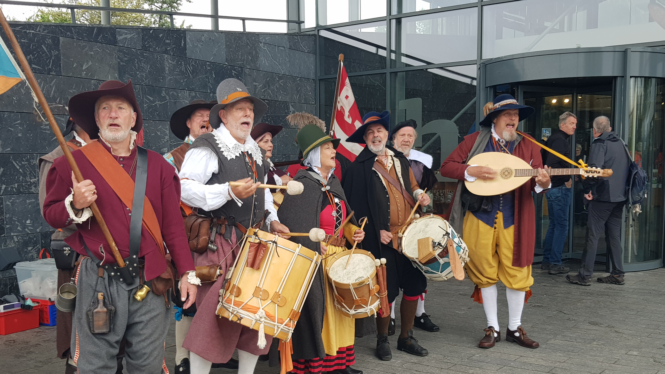 Mensen in historische kostuums spelen muziekinstrumenten, waaronder trommels en een luit, bij een glazen gebouw.