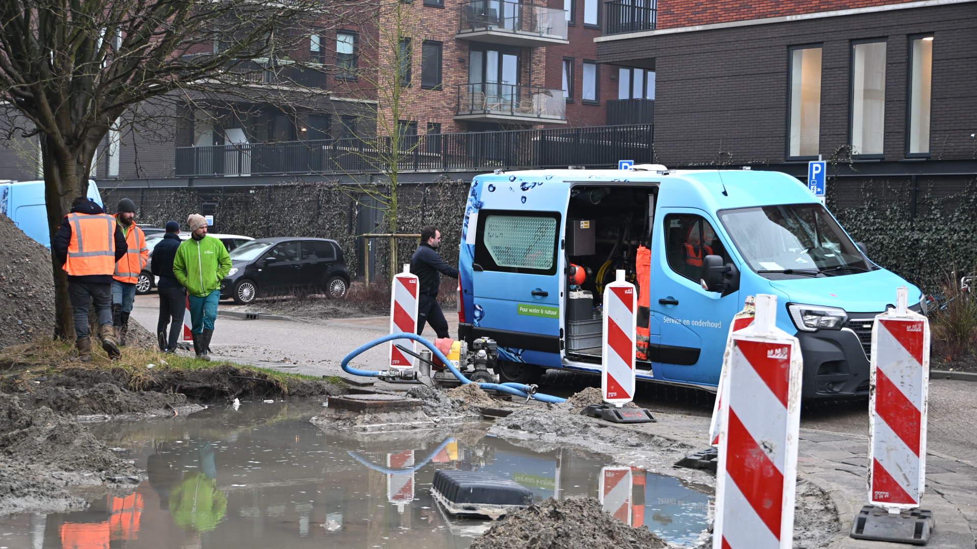 Bouwvakkers werken naast een blauw servicevoertuig rond een modderige waterplas, omgeven door waarschuwingsborden.