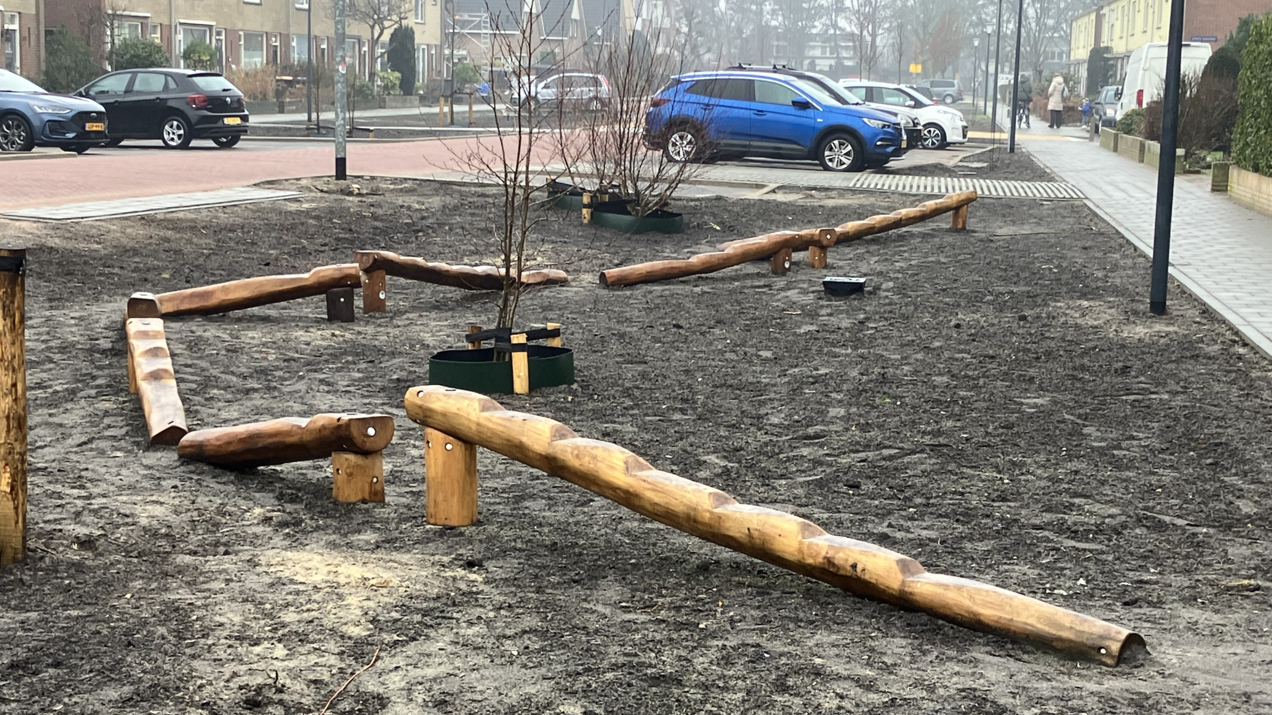 Houten speeltoestellen op een modderige speelplaats, met jonge boompjes en geparkeerde auto's op de achtergrond.