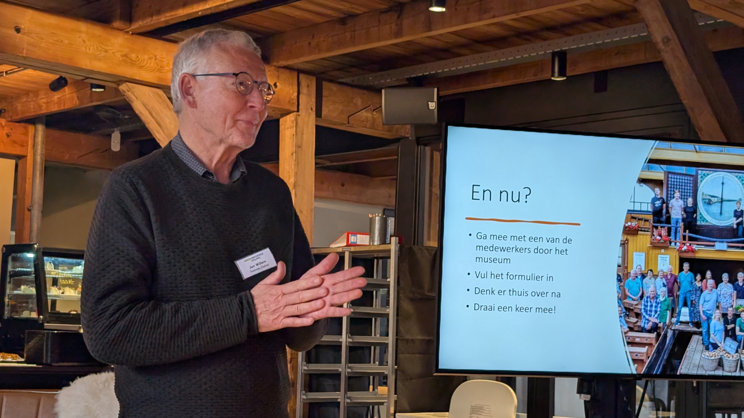 Een man spreekt voor een scherm met tekst over museumactiviteiten, in een ruimte met houten balken.