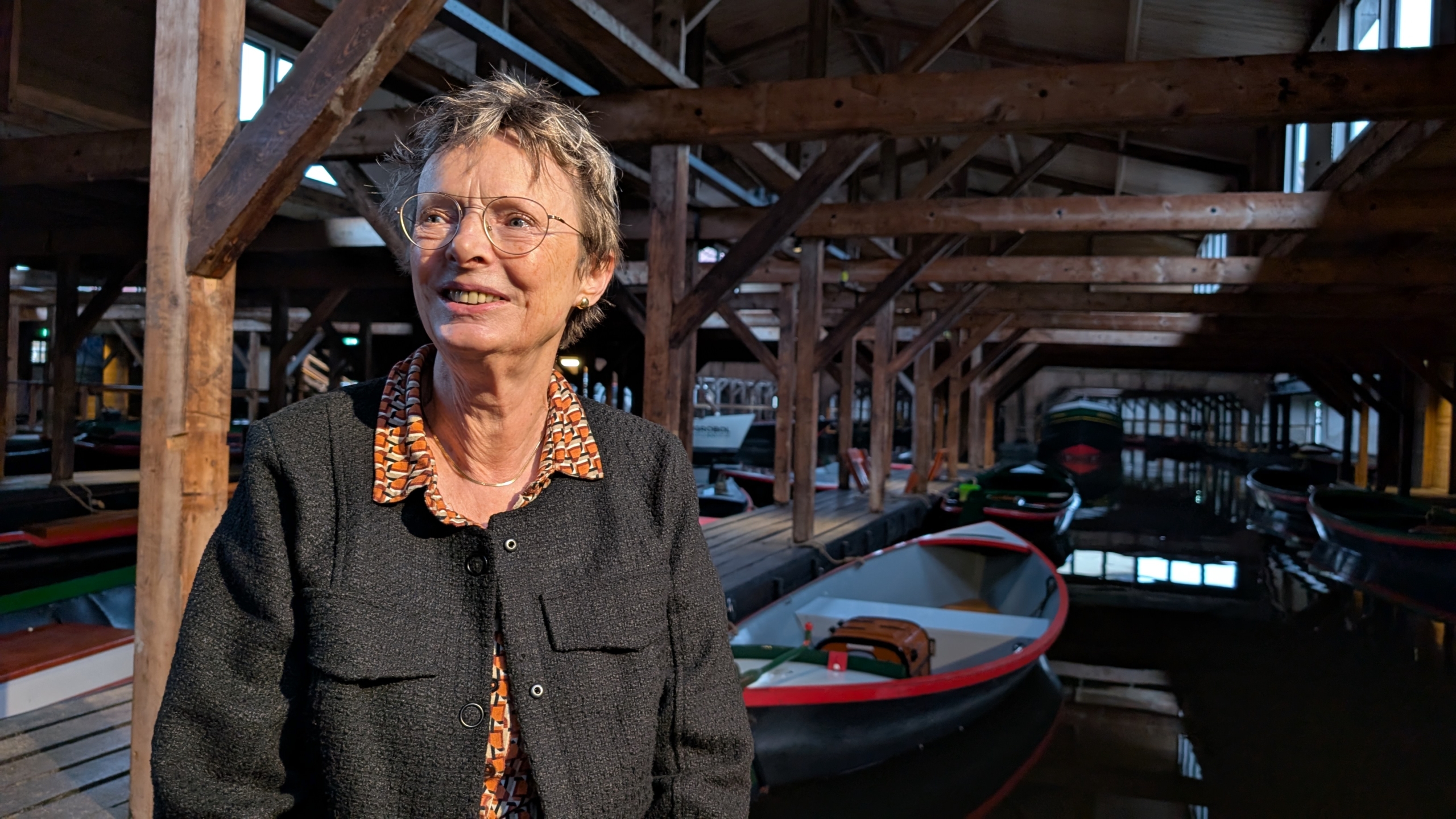 Vrouw met bril en grijs haar in een houten scheepswerf, omringd door boten.