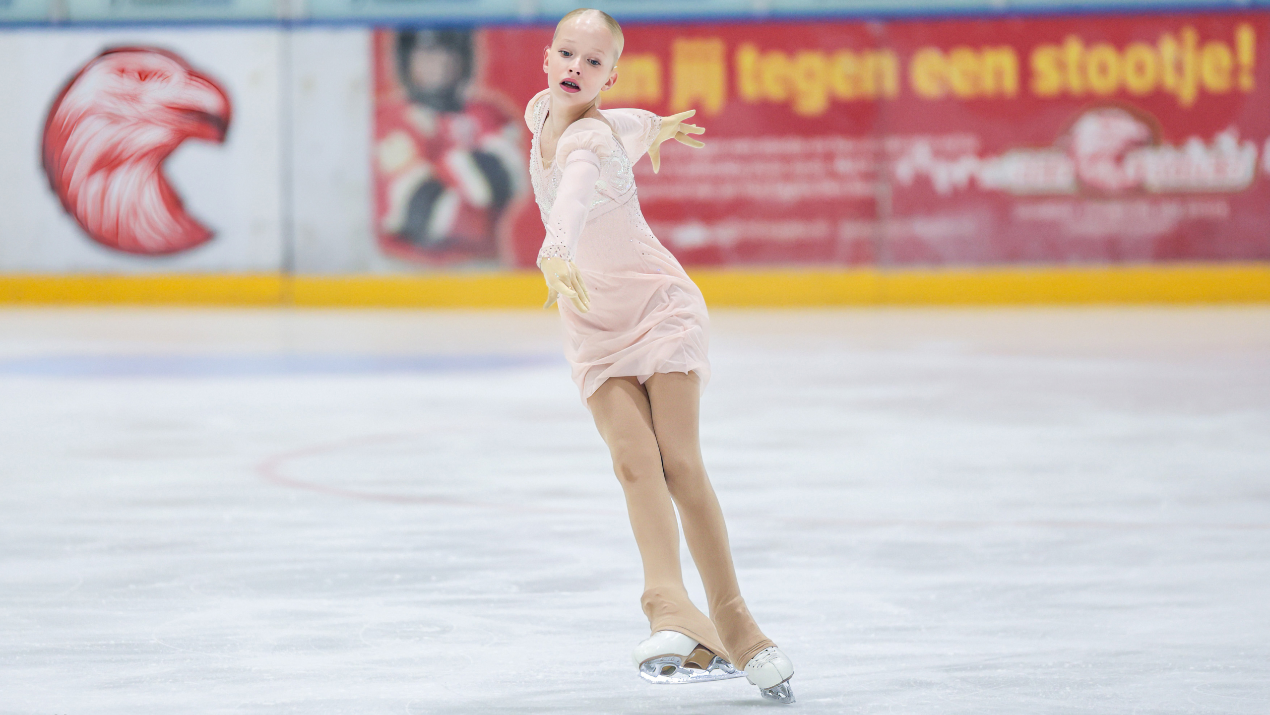 Een kunstschaatsster in een roze jurk doet een sierlijke beweging op de ijsbaan.