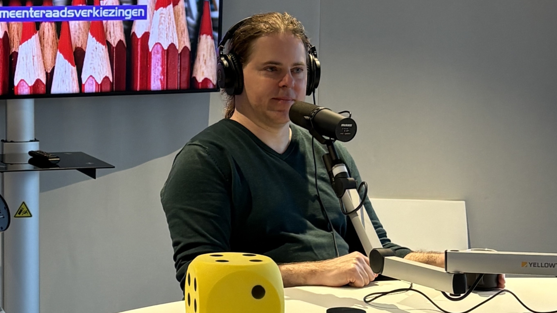 Een man met een koptelefoon zit voor een microfoon in een opnamestudio met een grote gele dobbelsteen op tafel.