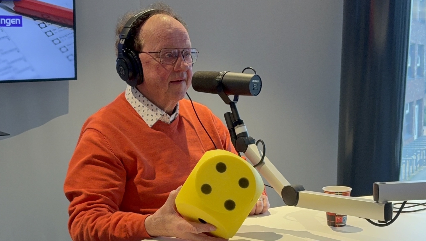 Man in een oranje trui met een koptelefoon houdt een grote gele dobbelsteen vast in een radiostudio.