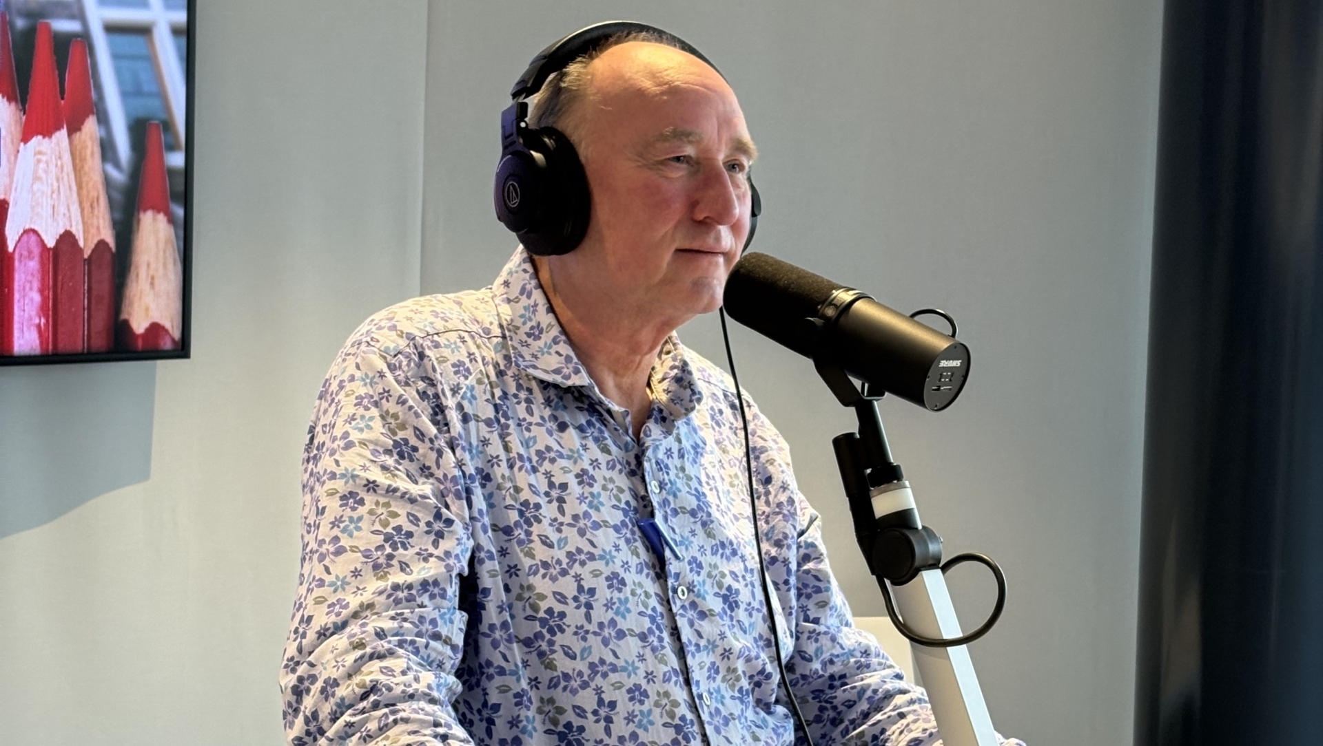 Man met een koptelefoon en een blauwe bloemenblouse spreekt in een microfoon in een studio.