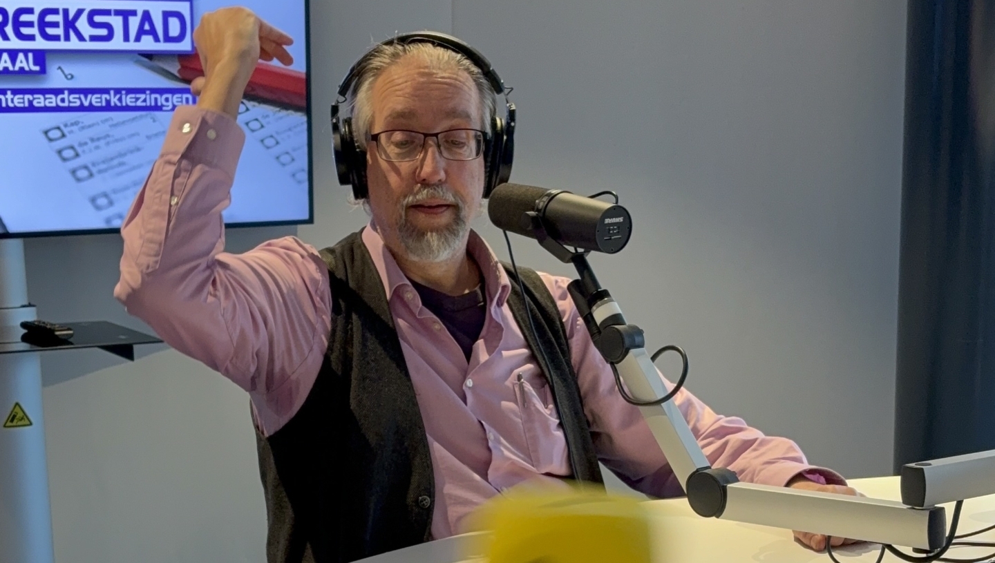 Man met hoofdtelefoon gooit een gele dobbelsteen aan een radiostudio tafel. Op de achtergrond hangt een scherm.