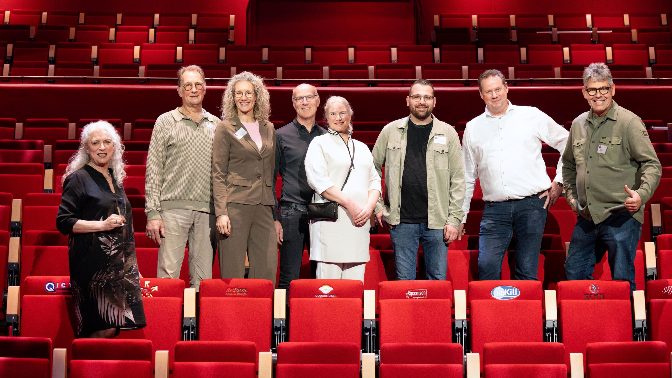 Een groep van acht mensen poseert voor een rij lege rode theaterstoelen.