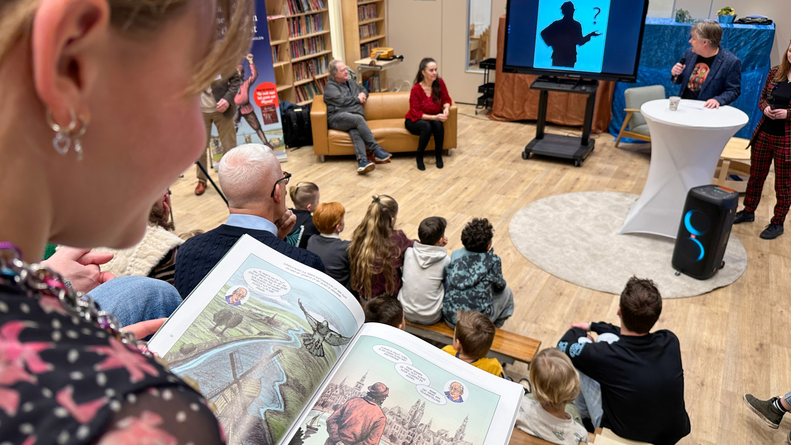 Een meisje leest een stripboek terwijl een presentatie met een projectiescherm plaatsvindt in een bibliotheek.