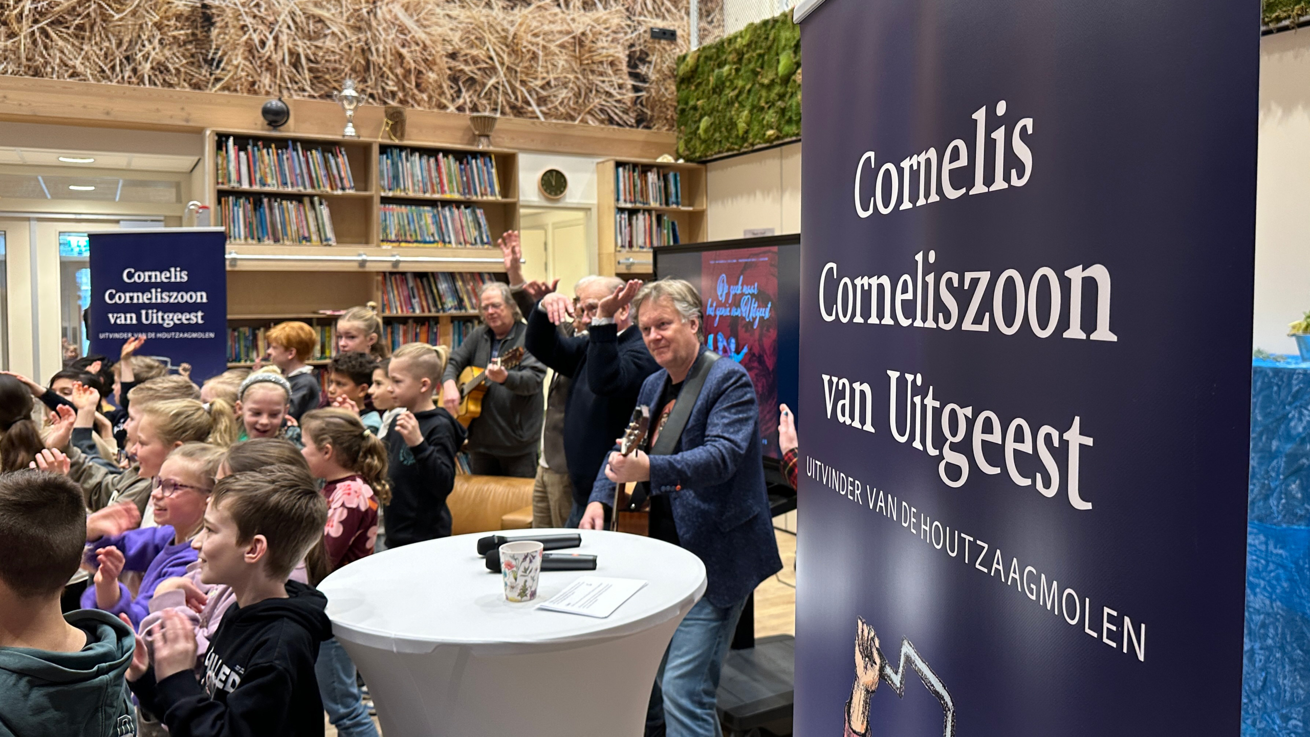 Kinderen en volwassenen in een bibliotheek applaudisseren tijdens een muzikaal optreden, met banners over Cornelis Corneliszoon van Uitgeest.
