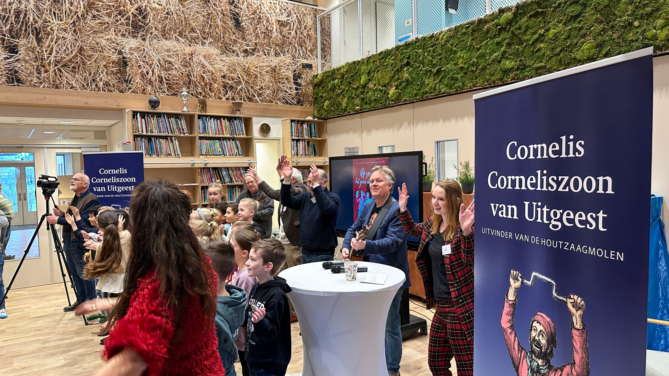 Een groep kinderen en volwassenen klapt en zingt in een bibliotheek, naast een bord over Cornelis Corneliszoon van Uitgeest.