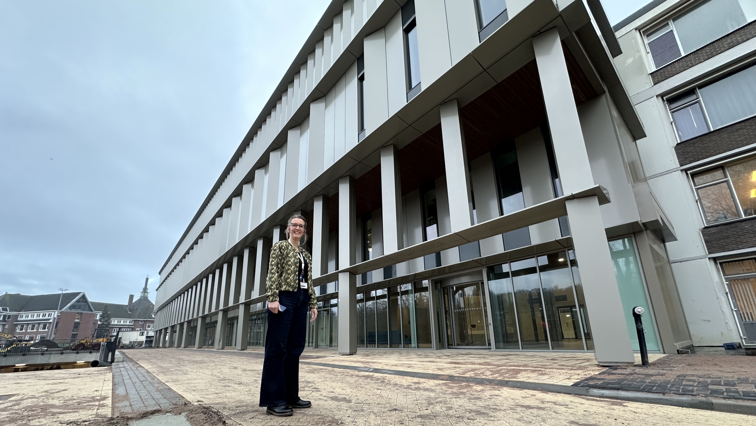 Vrouw staat voor een modern gebouw met grote ramen en pilaren op een bewolkte dag.