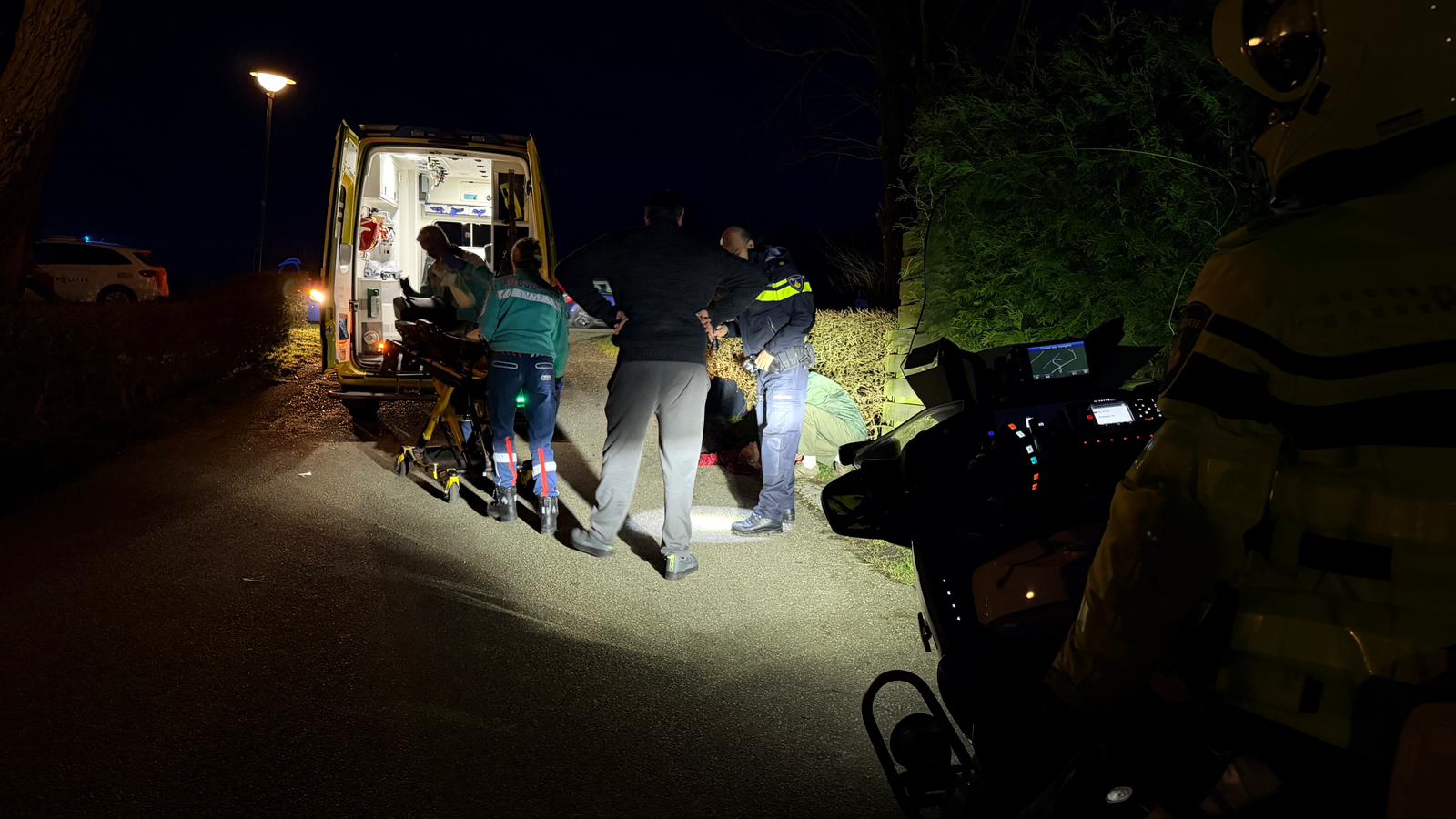 Ambulancepersoneel en politie zijn 's nachts bezig bij een draaiende ambulance, met een paar mensen op de grond.