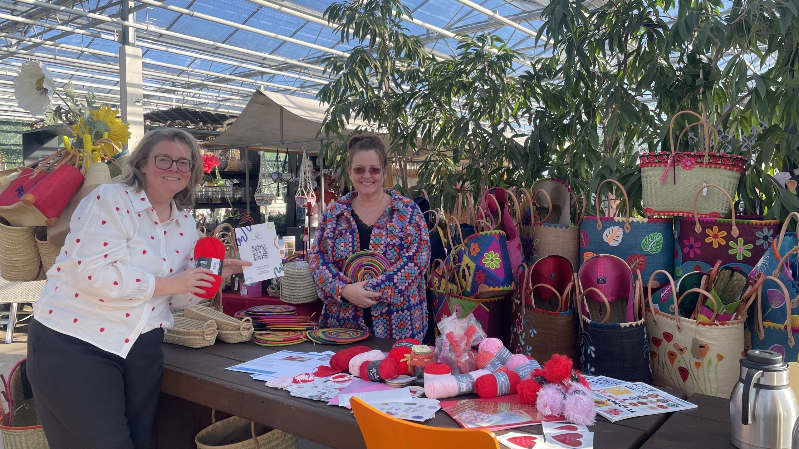 Twee vrouwen glimlachen achter een tafel vol kleurrijke manden, garen en decoraties in een kas met planten.