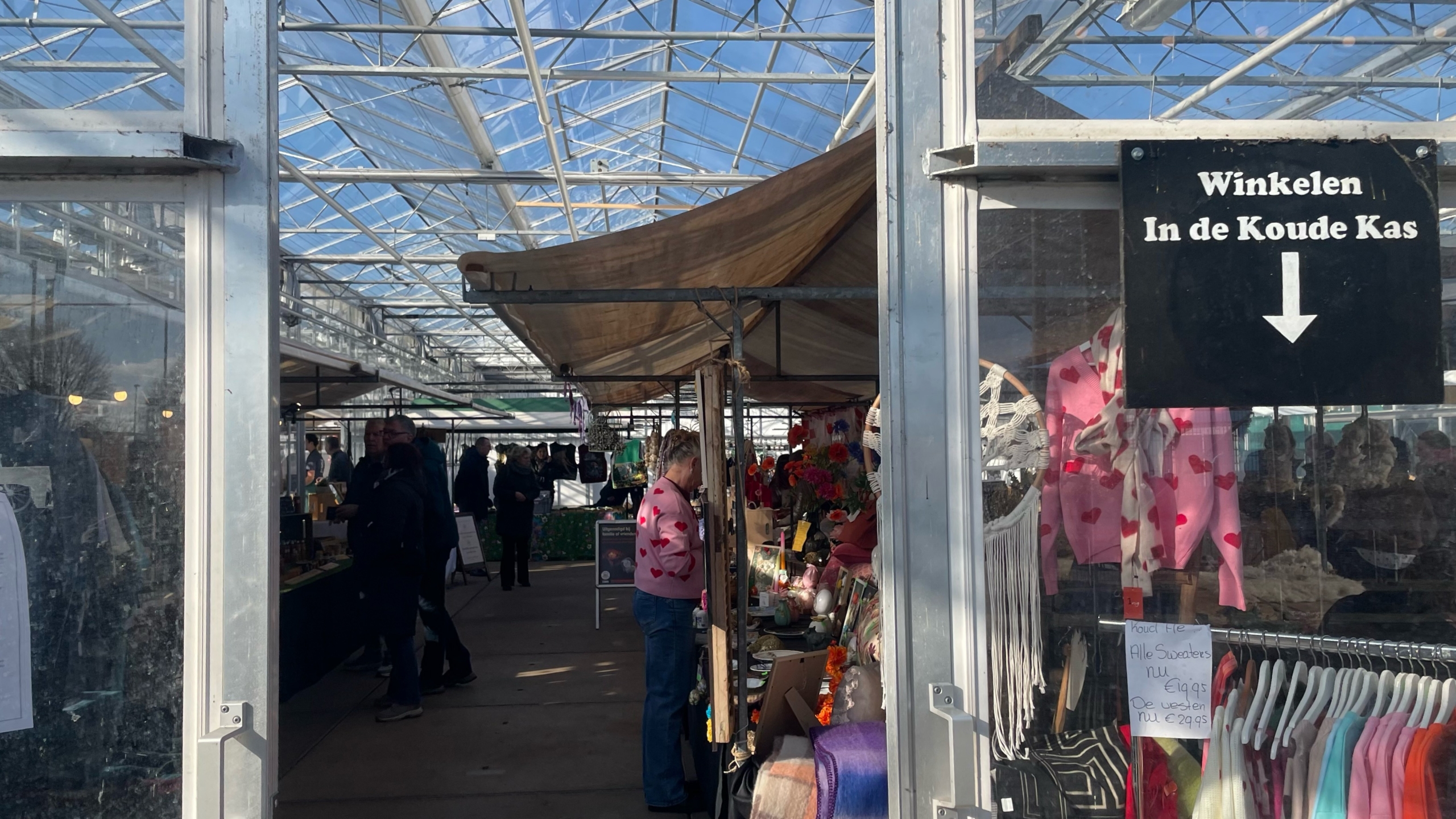 Mensen winkelen op een binnenmarkt onder een glazen dak met bord "Winkelen In de Koude Kas" en kleurrijke kleding te koop.