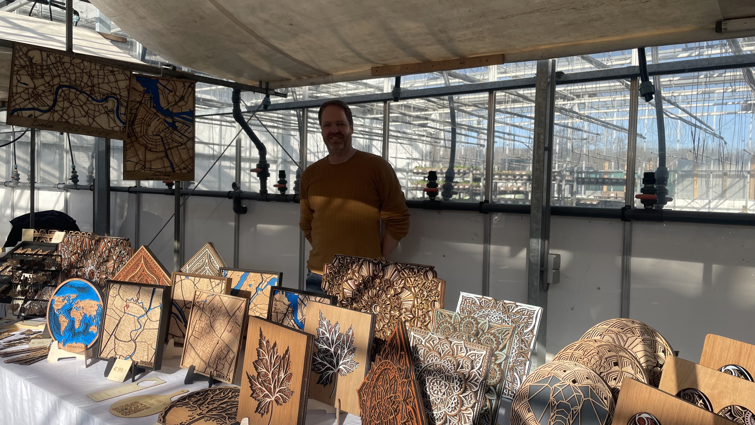 Man bij een kraampje met gedetailleerd houtsnijwerk en kaarten, binnen in een kas met glazen ramen.