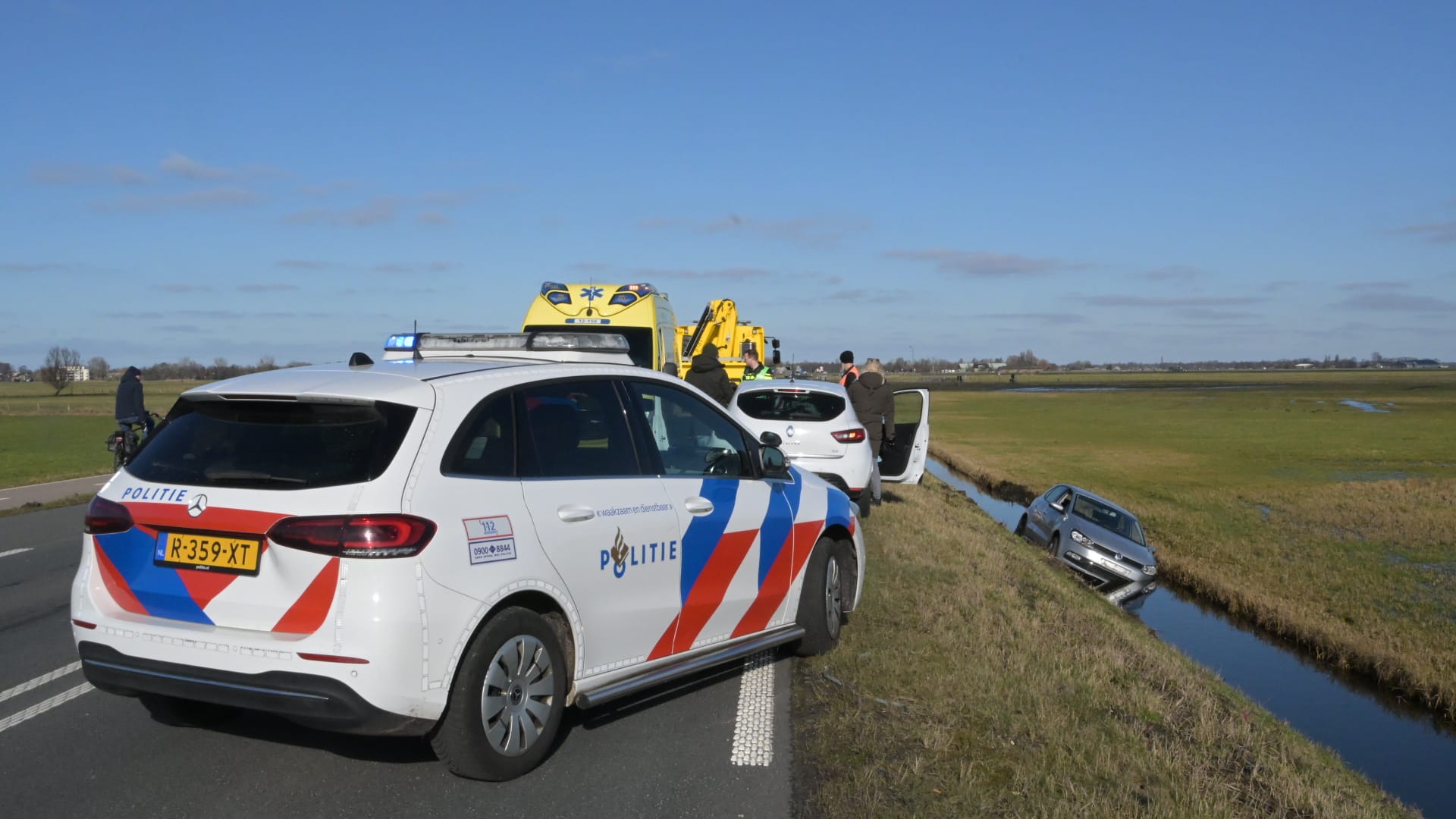 Politieauto en ambulance bij een ongeluk met een auto in een sloot langs een weg in een landelijk gebied.