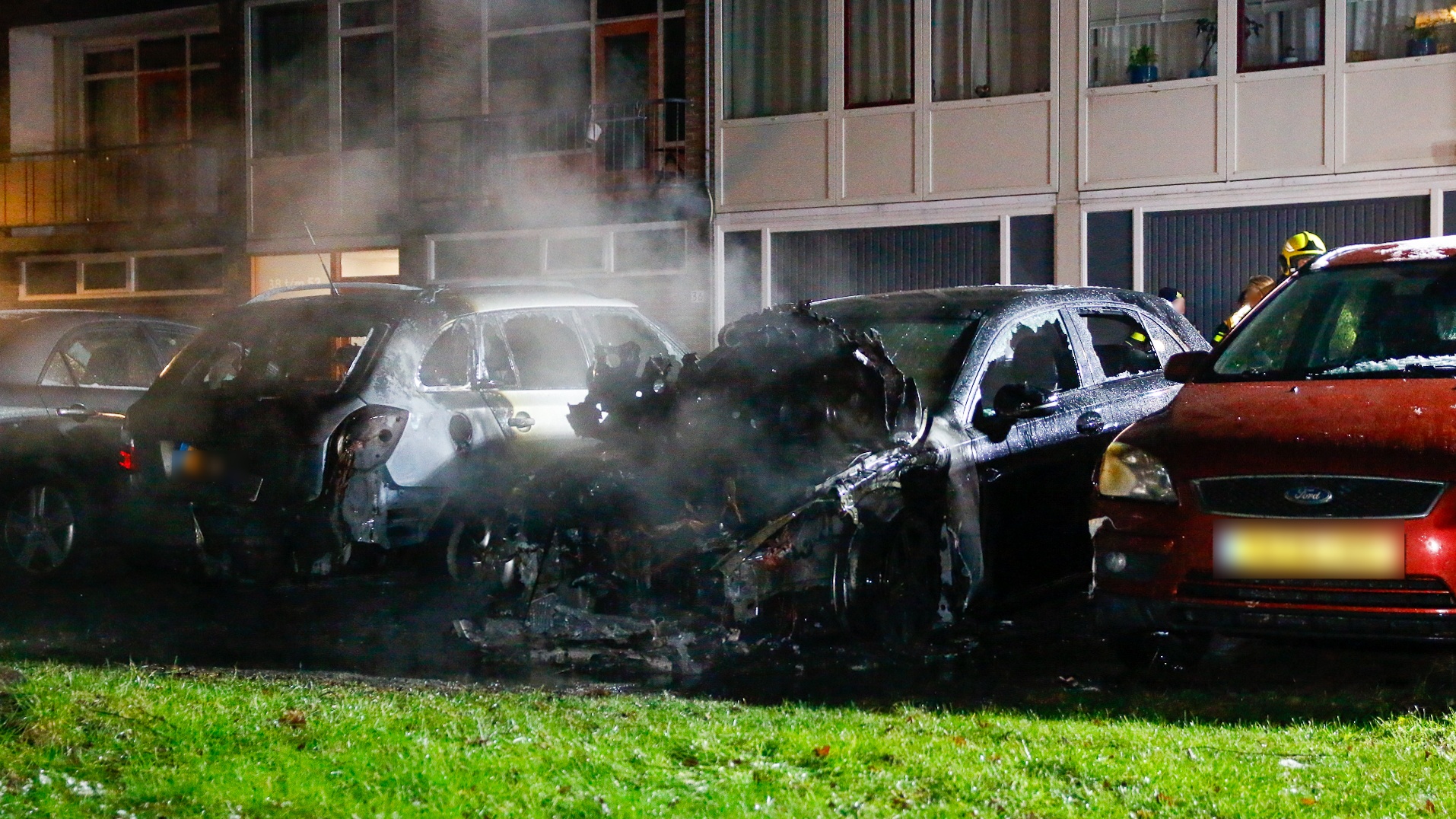 Drie auto's, waarvan één zwaar beschadigd door brand, met een brandweerman op de achtergrond.