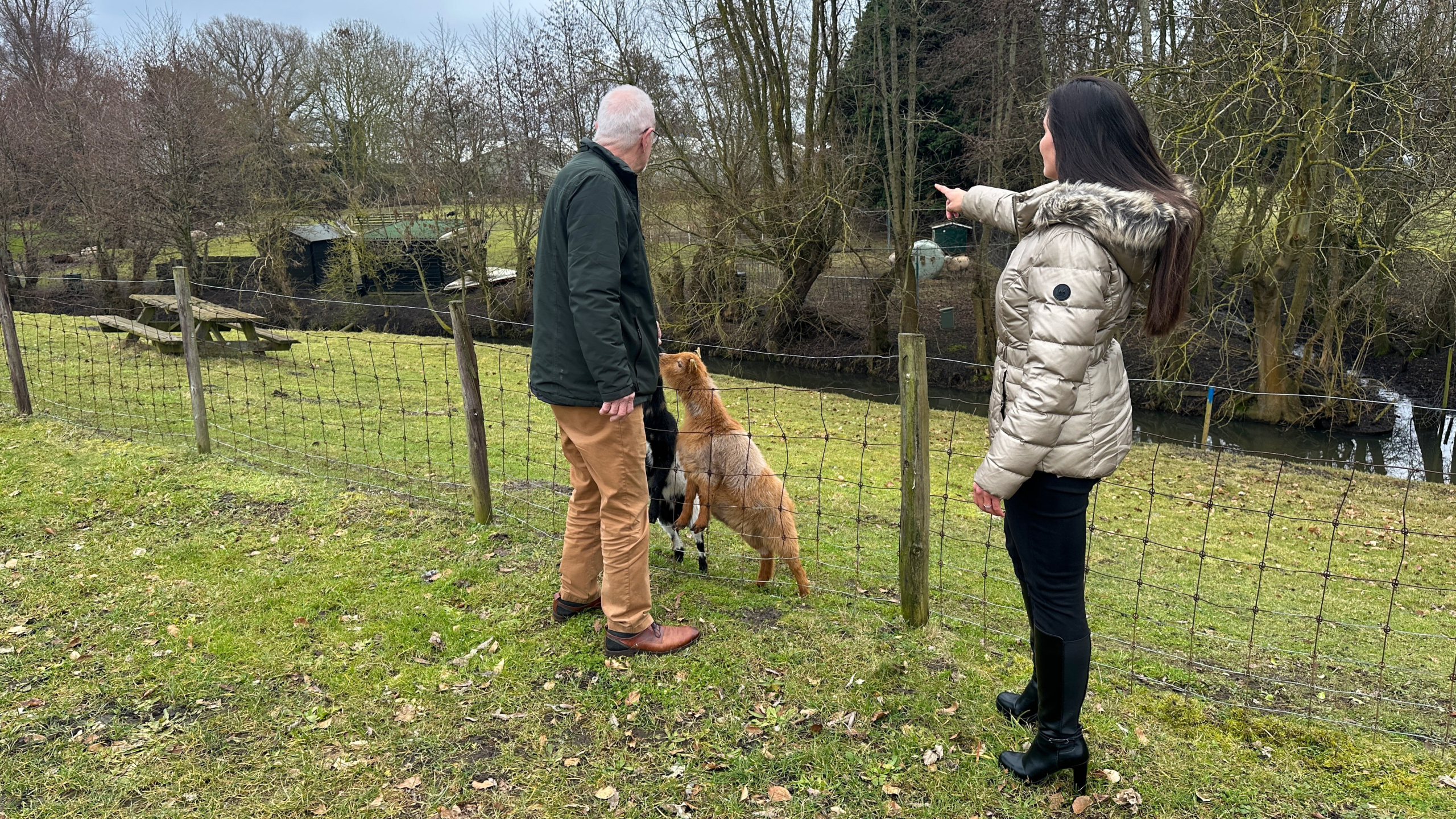 Twee mensen en twee geiten staan bij een hek, terwijl een vrouw met jas ergens naartoe wijst in een landelijke omgeving.