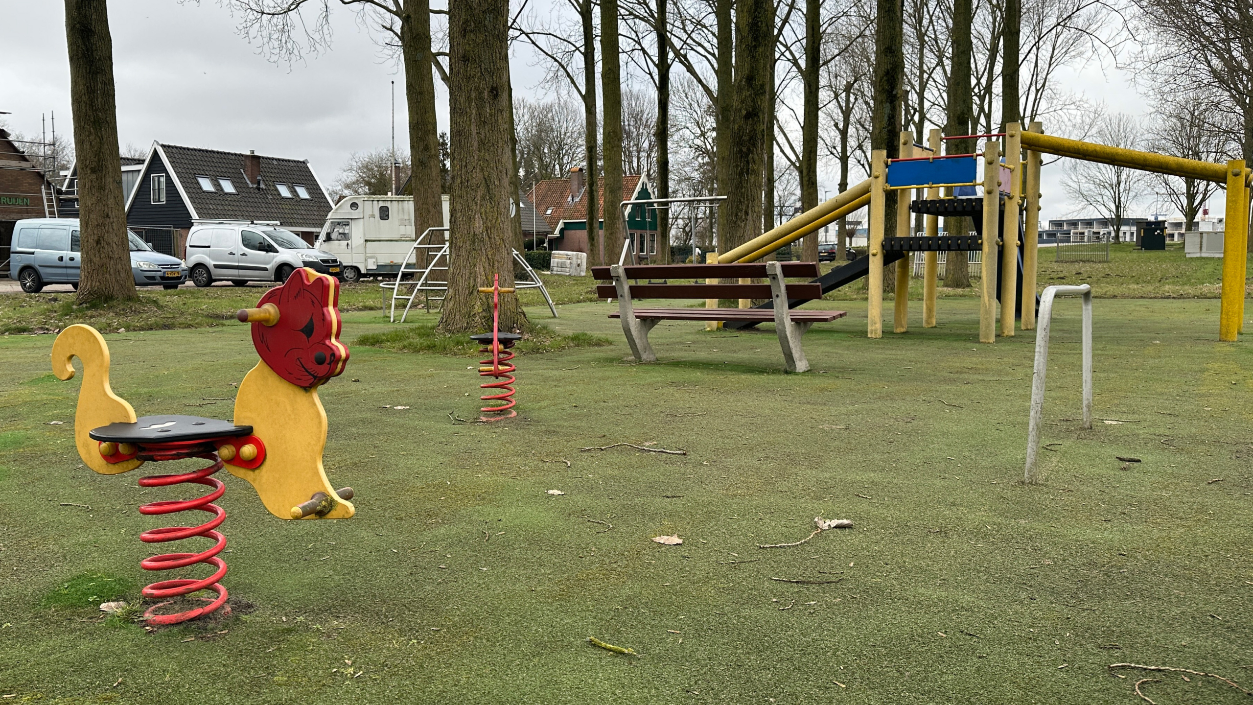 Speeltuin met speeltoestellen zoals een wipkip in de vorm van een kat, glijbaan, en een bankje, omgeven door bomen.