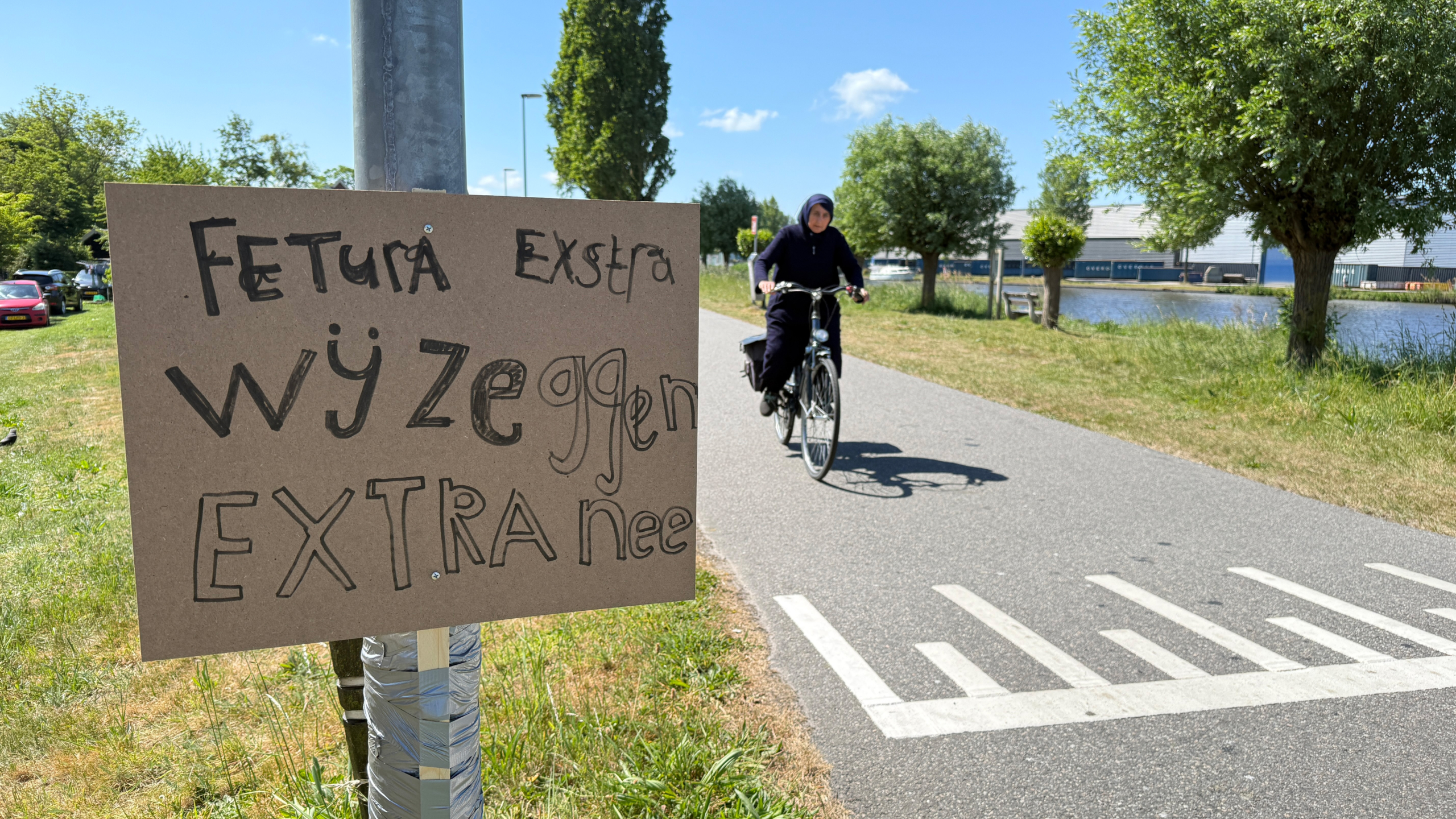 Een vrouw fietst op een pad langs een waterkant, met een bord voorop met de tekst "Extra wijzen extra nee".
