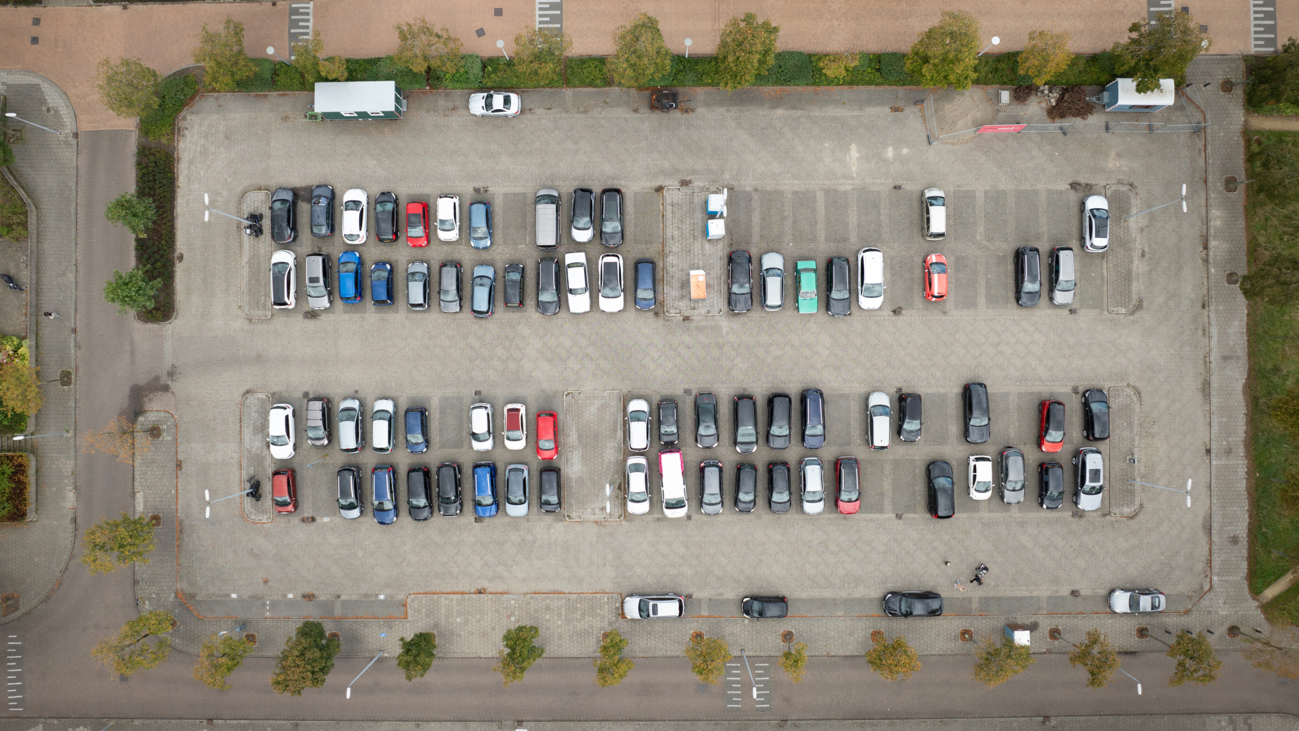 Luchtfoto van een vol parkeerterrein met diverse gekleurde auto's, omringd door bomen en wegen.