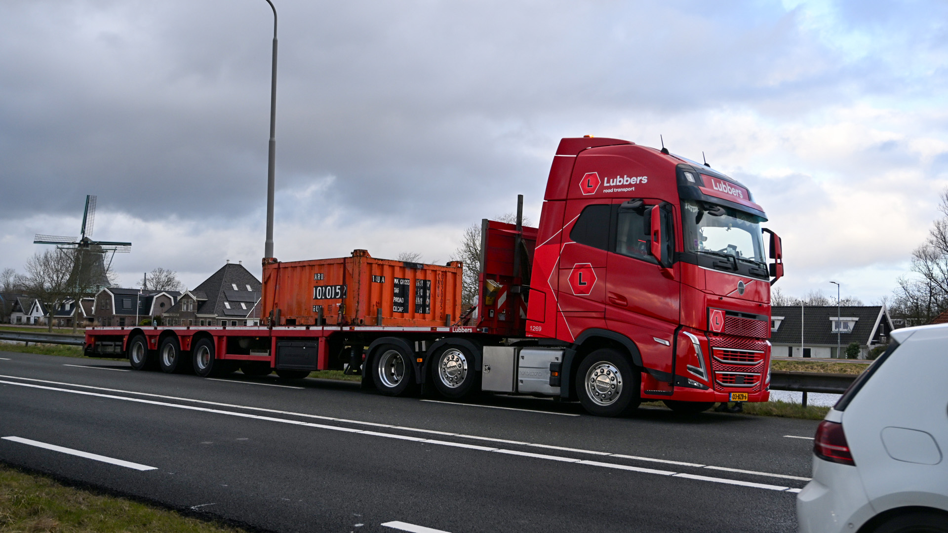 Rode vrachtwagen van Lubbers met lange oplegger en container, geparkeerd langs een weg met gebouwen en een molen op de achtergrond.