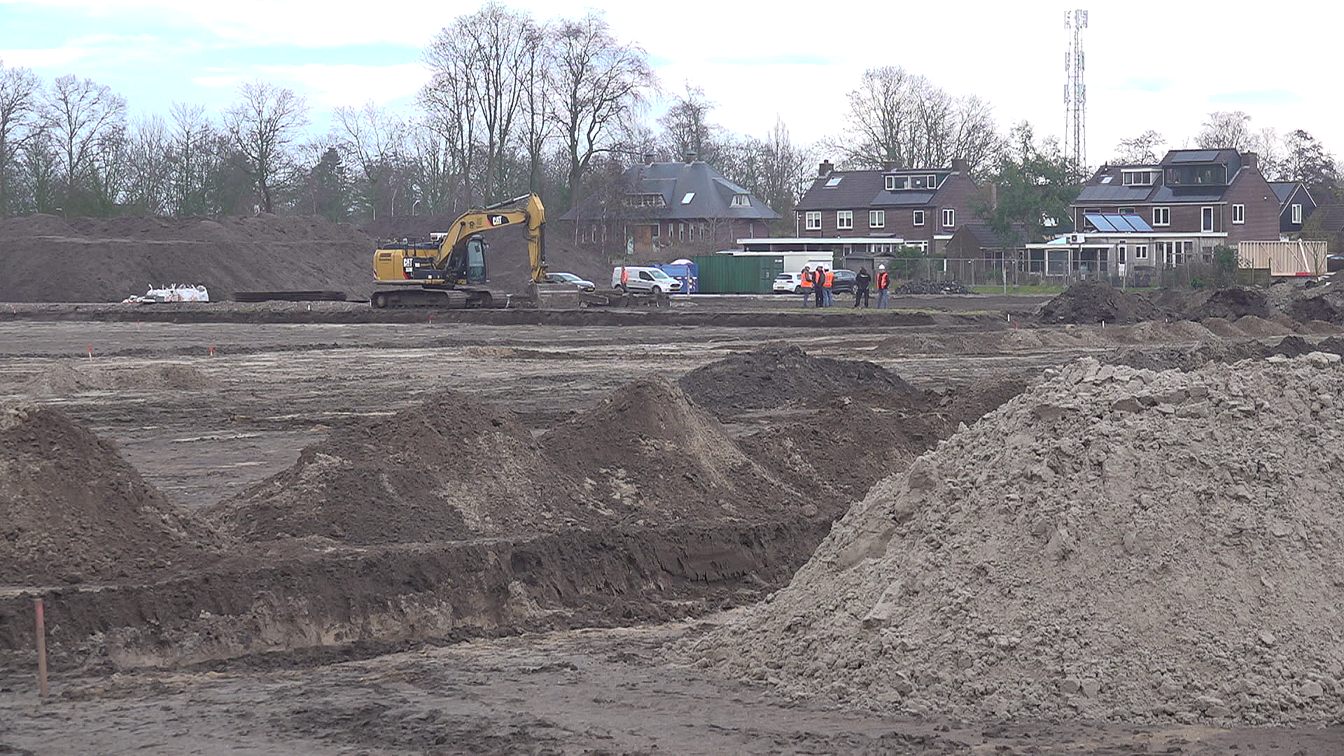 Graafmachine en werklieden op een bouwterrein met heuvels aarde, omringd door bomen en huizen.