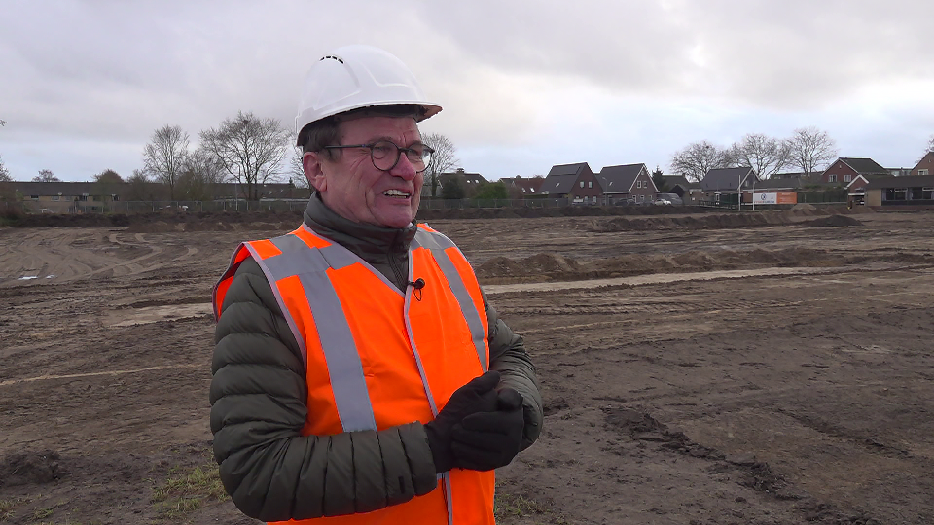 Man in een oranje veiligheidsvest en witte helm op een bouwterrein met huizen in de achtergrond.