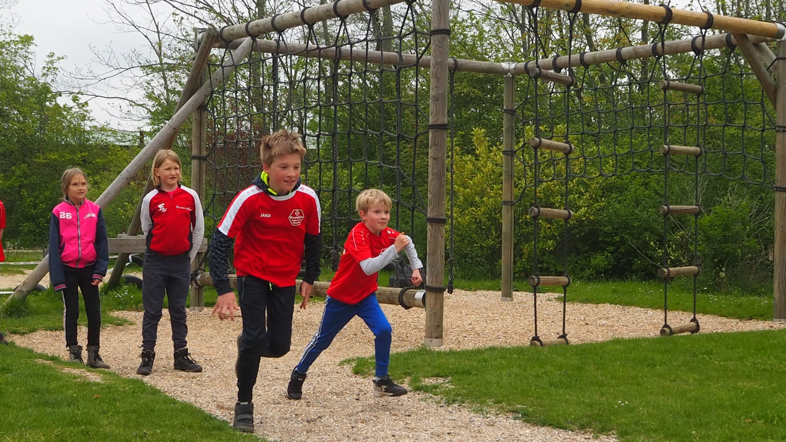 Kinderen in sportkleding rennen bij een klimrek in een speeltuin, met bomen op de achtergrond.