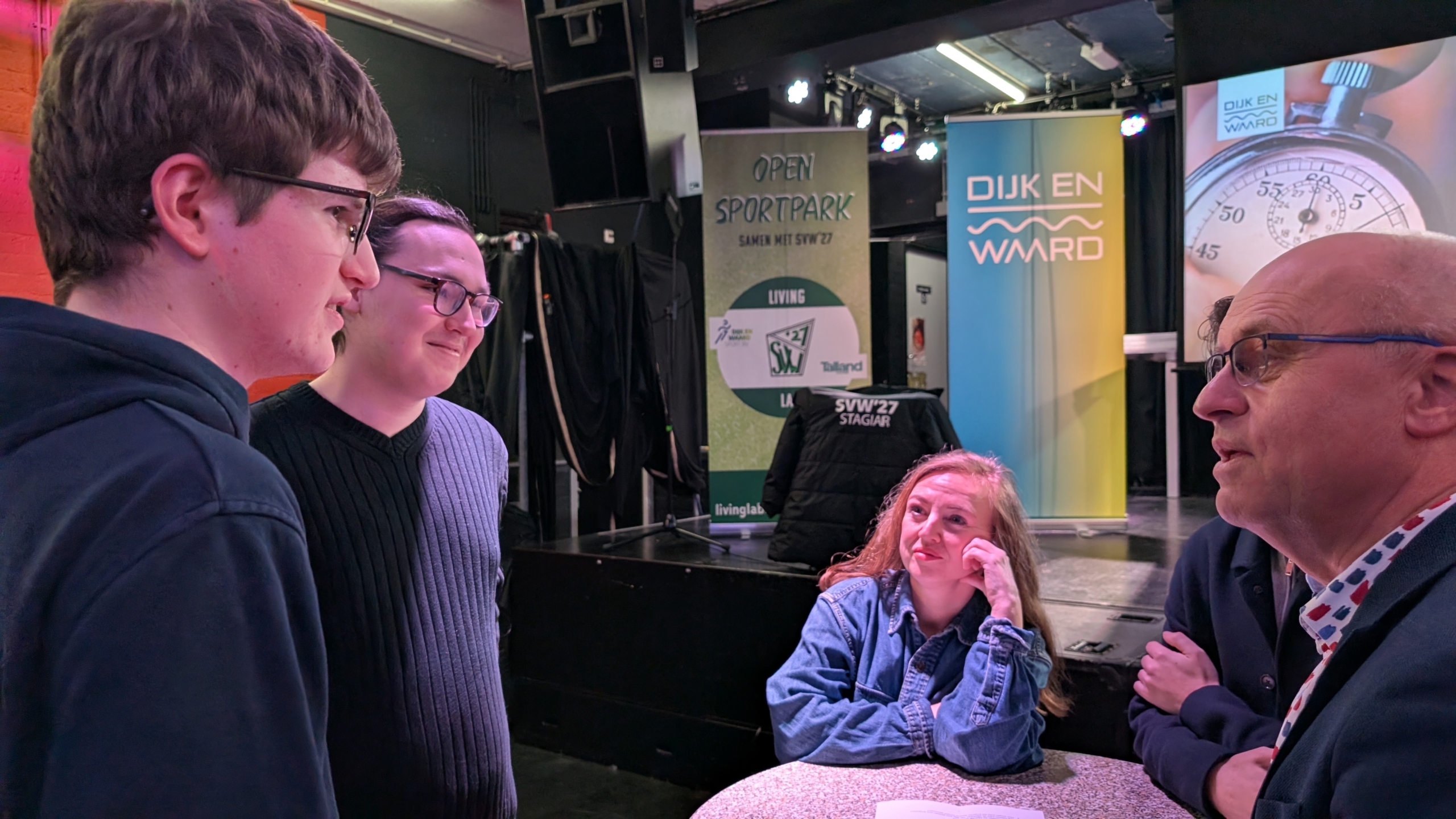 Vier mensen zijn in gesprek rondom een tafel in een zaal met banners van "Open Sportpark" en "Dijk en Waard" op de achtergrond.
