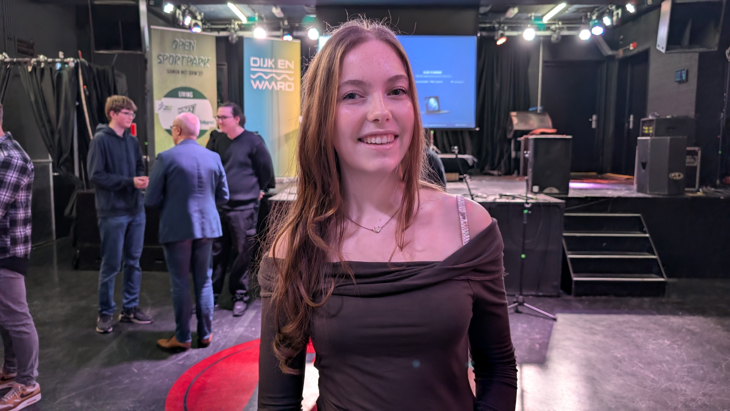 Jonge vrouw glimlacht naar de camera in een evenementhal met mensen en schermen op de achtergrond.